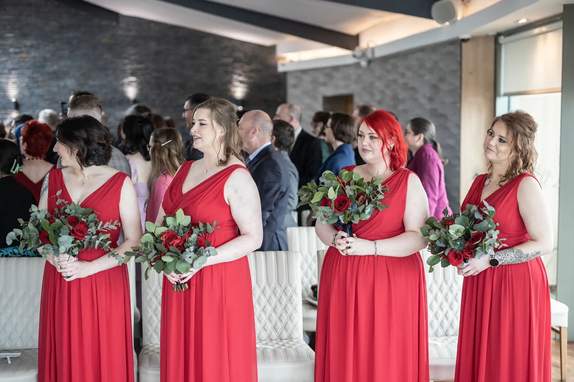 Four bridesmaids in matching sleeveless red dresses hold bouquets with red and white flowers and greenery. They stand in a bright room with cream chairs, a stone feature wall, and guests in the background, some seated and some standing.