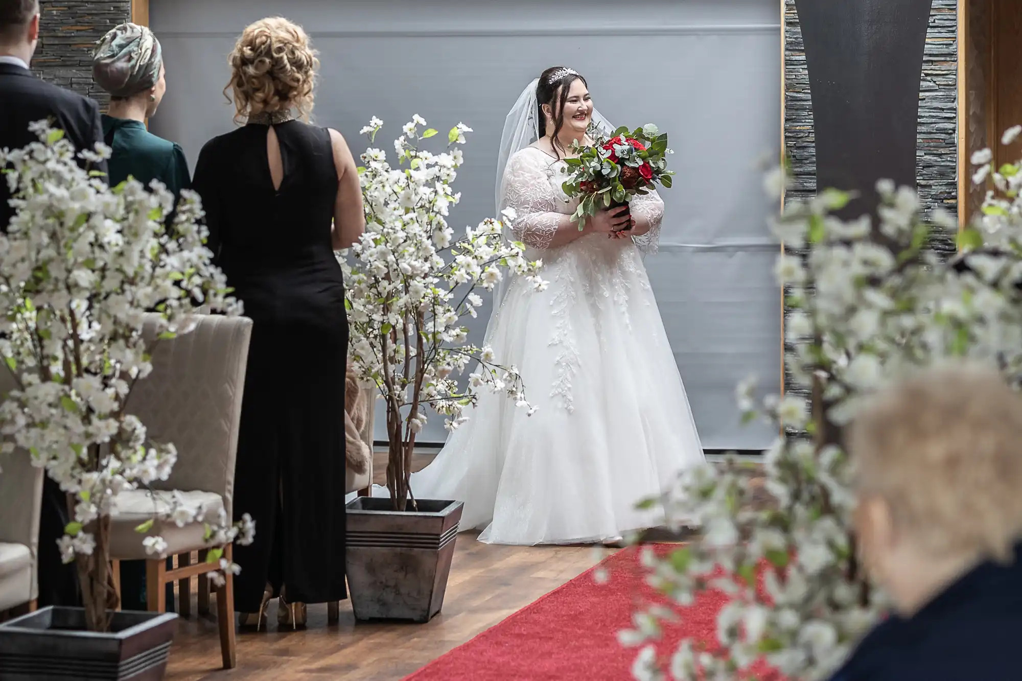 A bride in a white wedding dress and veil, holding a bouquet of red and white flowers, stands smiling at the start of an indoor aisle decorated with white flowering branches. Guests stand and sit along the aisle, which is covered by a red carpet.