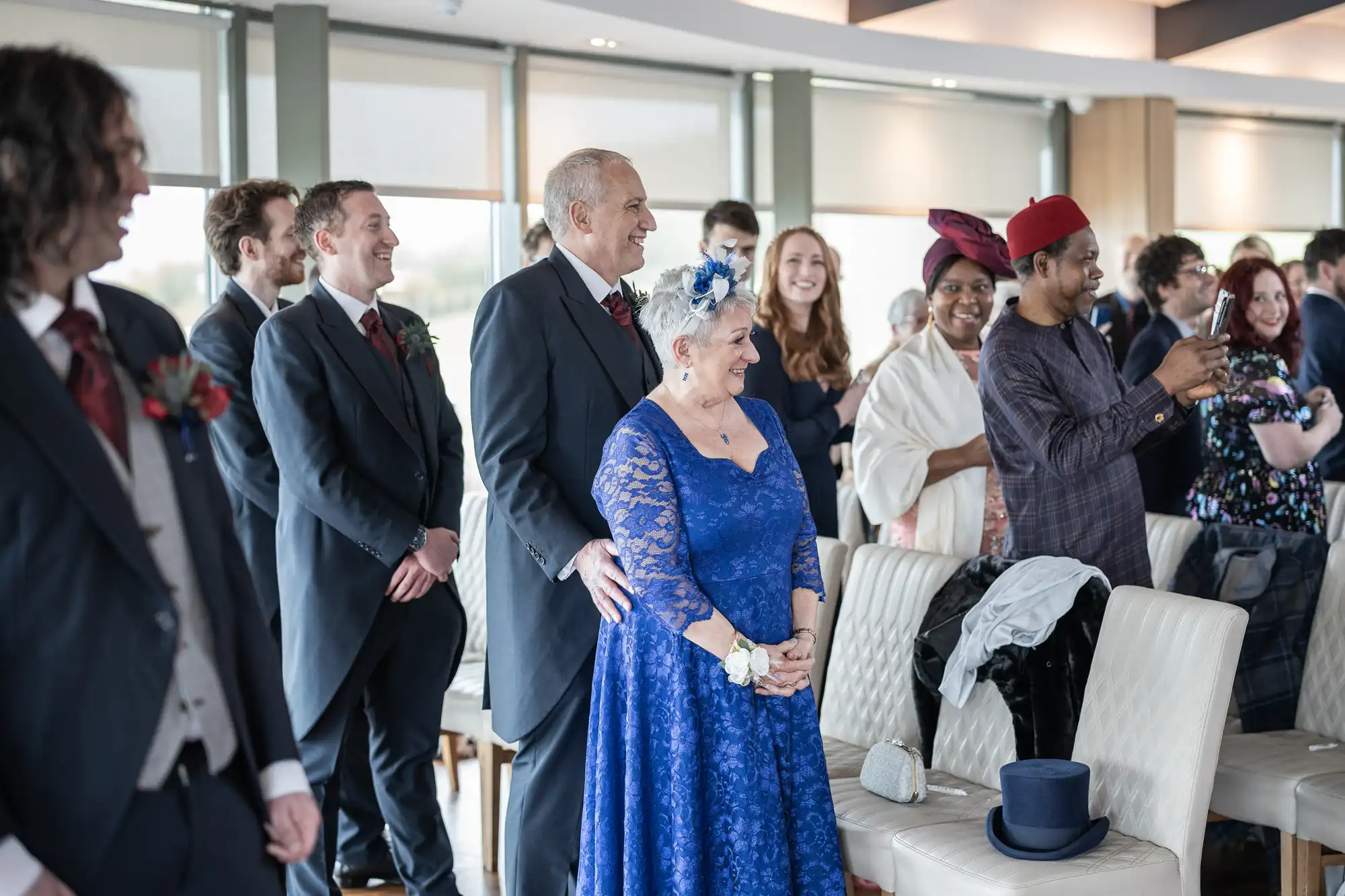 A group of people dressed formally at an indoor event, possibly a wedding. A smiling woman in a blue lace dress stands beside a suited man. Guests in suits and colourful dresses are seated and standing, some smiling, others watching or taking photos.
