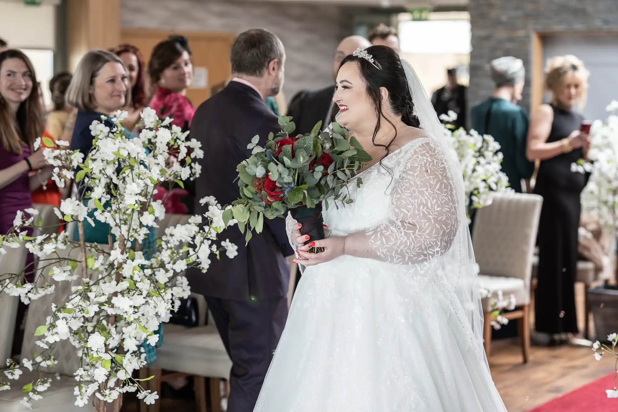 A bride in a white lace wedding dress and veil holds a bouquet of red and green flowers, smiling as she walks down an aisle lined with white floral arrangements. Guests in formal attire stand and watch, some smiling, in a bright indoor venue.