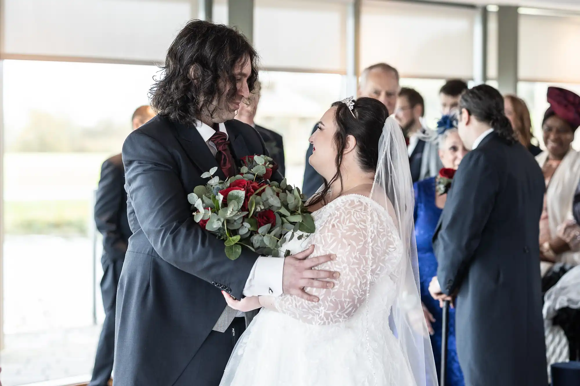 A groom with long curly hair and a dark suit embraces a smiling bride in a white dress and veil holding a bouquet of red roses. They face each other at an indoor wedding ceremony, surrounded by guests in formal attire.