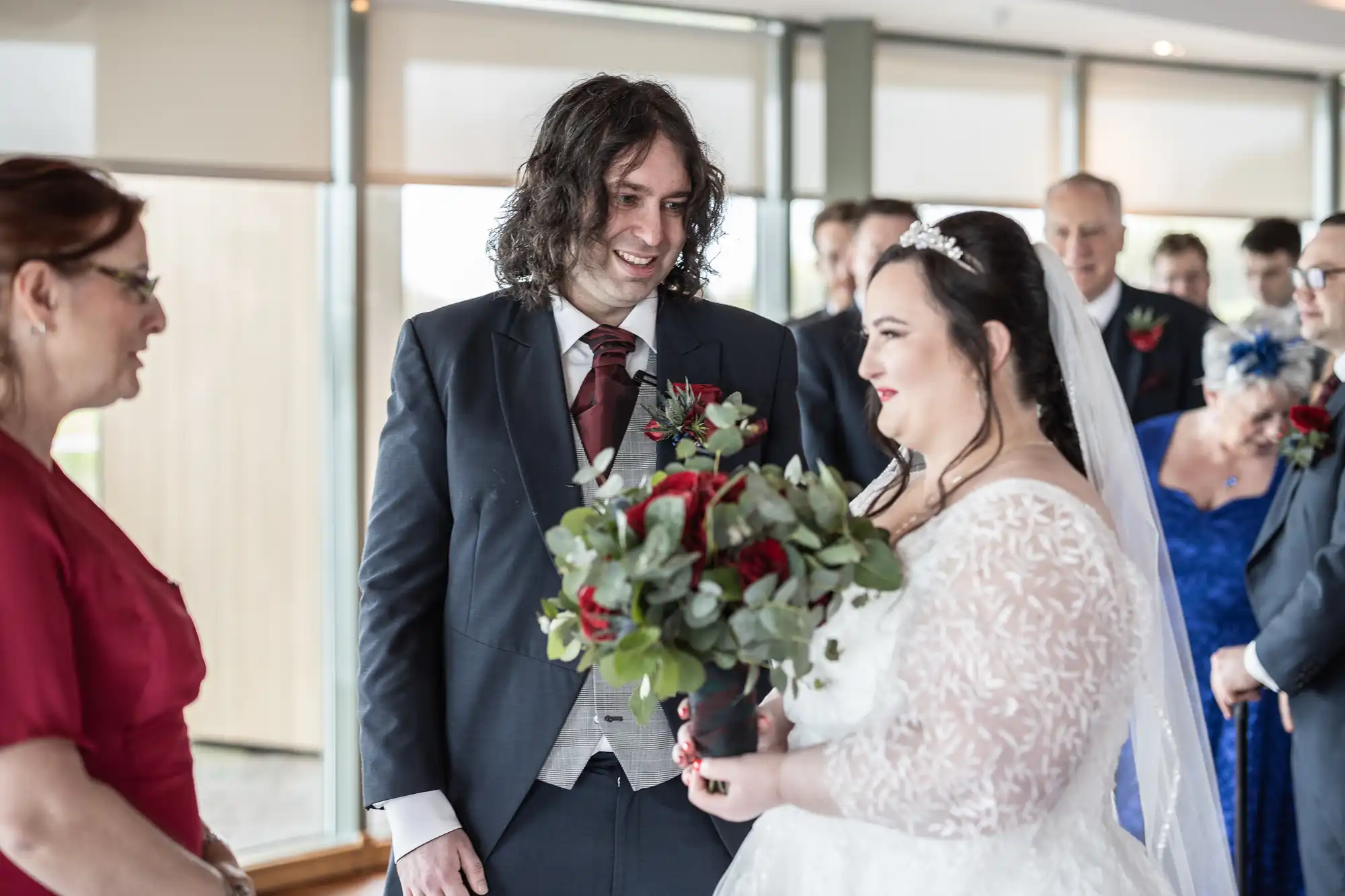 A bride in a white lace dress and veil holds a red and green bouquet, smiling at the groom in a dark suit and maroon tie. Guests, some dressed in blue, watch the ceremony indoors with large windows in the background.