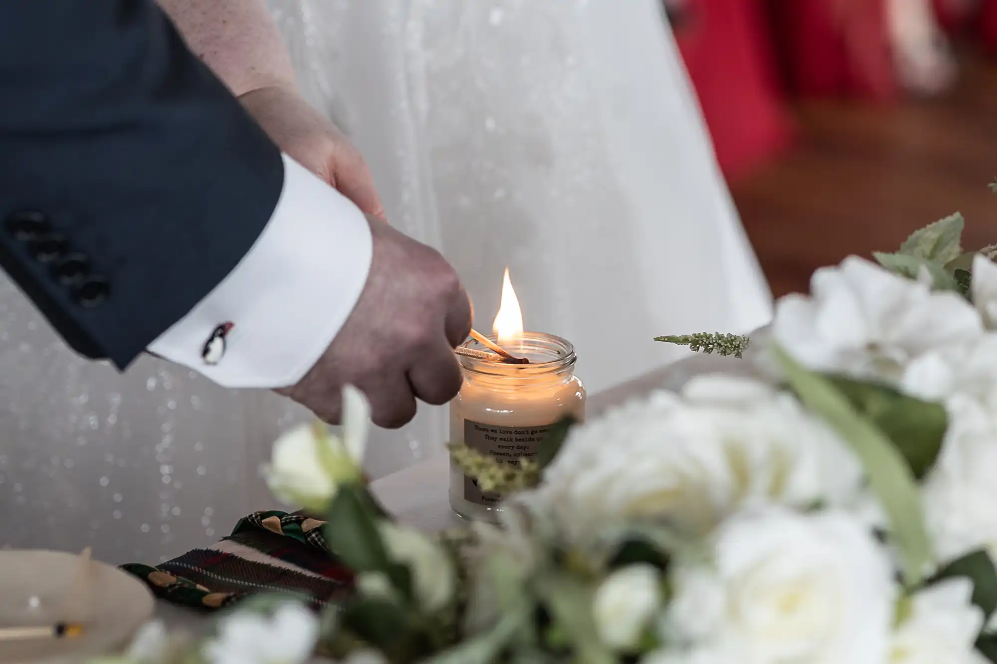 A close-up of two people lighting a candle together at a decorated table with white flowers. One person wears a dark suit and the other a white dress, suggesting a wedding or ceremonial event. The background is softly blurred.