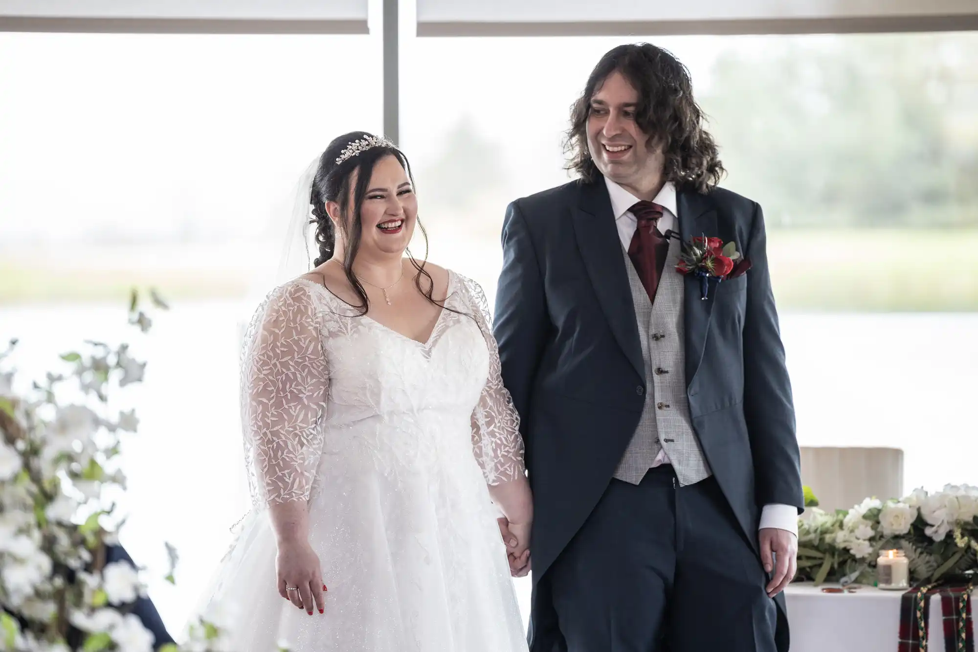 A bride in a white lace wedding dress and veil stands smiling, holding hands with a groom in a dark suit, grey waistcoat, and burgundy buttonhole. Both look happy, standing indoors near a window with white floral decorations nearby.