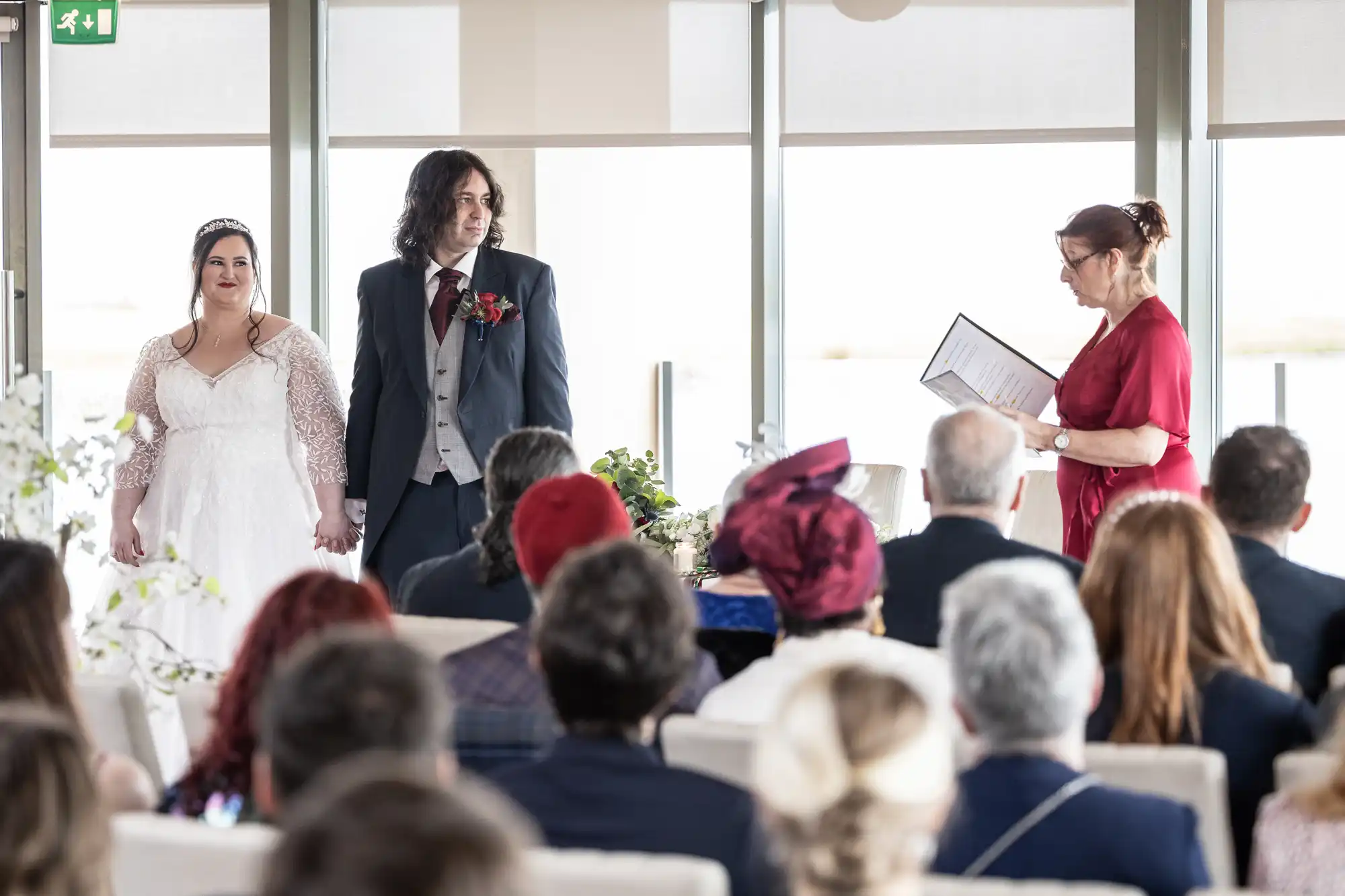 A bride in a white dress and a groom in a suit stand holding hands, facing a woman in a red dress holding papers, likely the celebrant. They are at the front of a room filled with seated wedding guests, some with hats, during a ceremony.