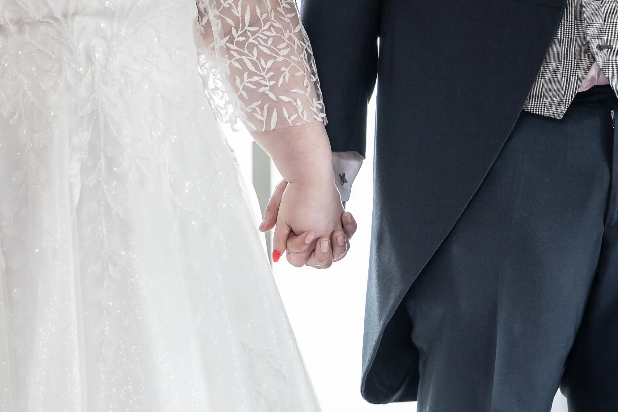 A close-up of a bride and groom holding hands. The bride wears a white lace-sleeved gown, and the groom wears a dark suit with a light checked waistcoat. Their hands are gently clasped, symbolising unity, against a bright, softly lit background.