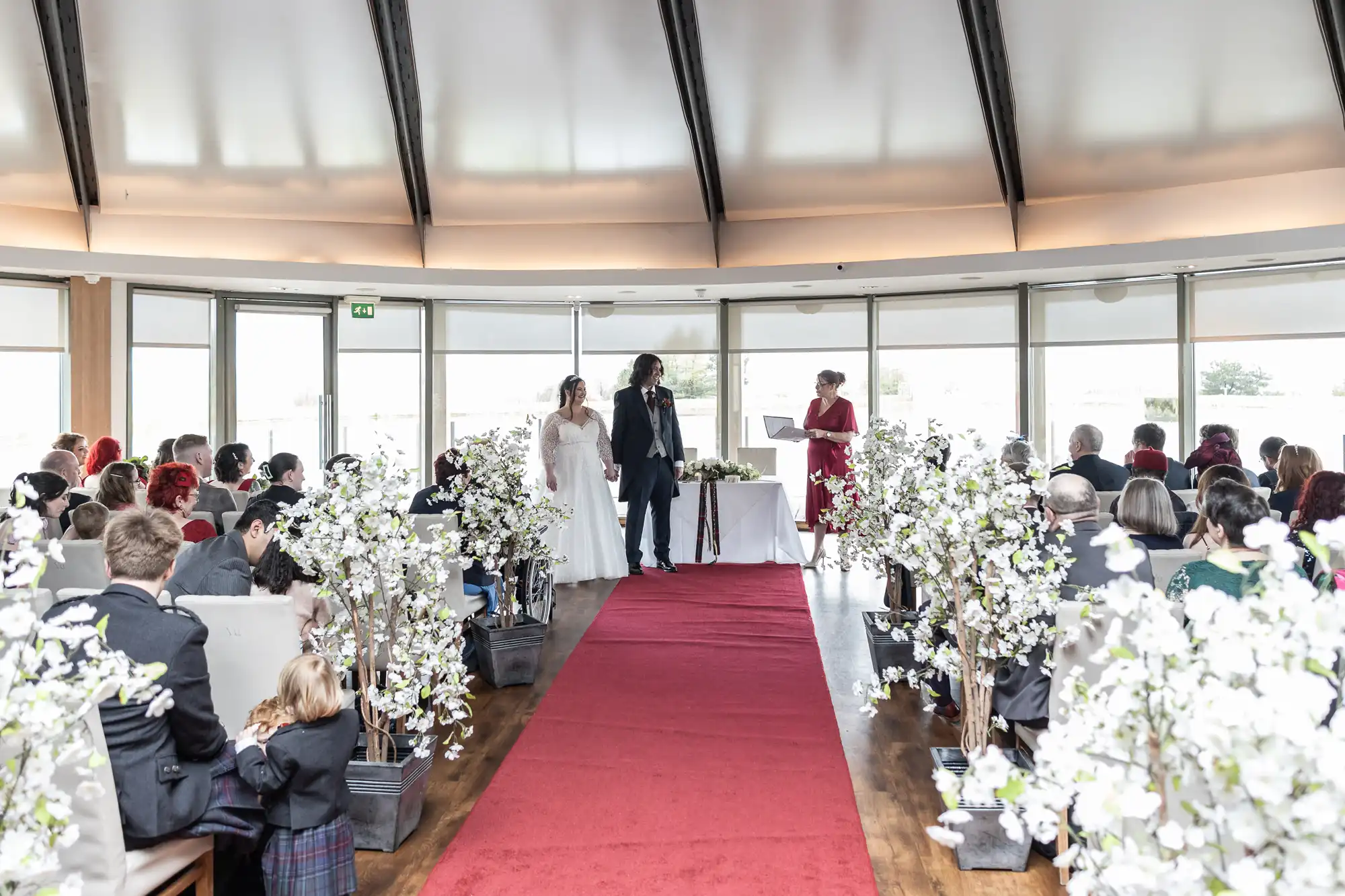A bride and groom stand at the altar with an officiant in a bright, modern venue. Guests are seated on both sides of a red aisle runner lined with white floral arrangements. Large windows curve around the room, letting in natural light.