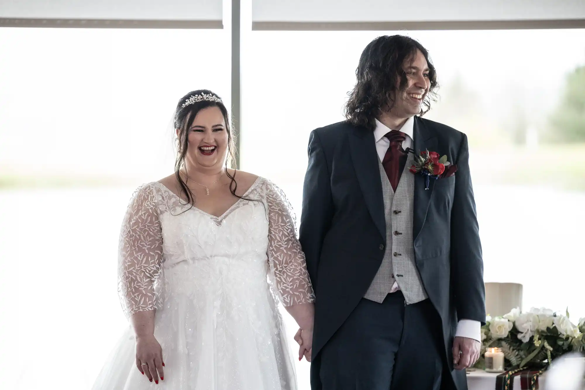 A smiling bride in a white lace wedding dress and tiara stands holding hands with a grinning groom in a dark suit, waistcoat, and burgundy tie. They are indoors, in front of large bright windows, with white flowers and candles on a table nearby.
