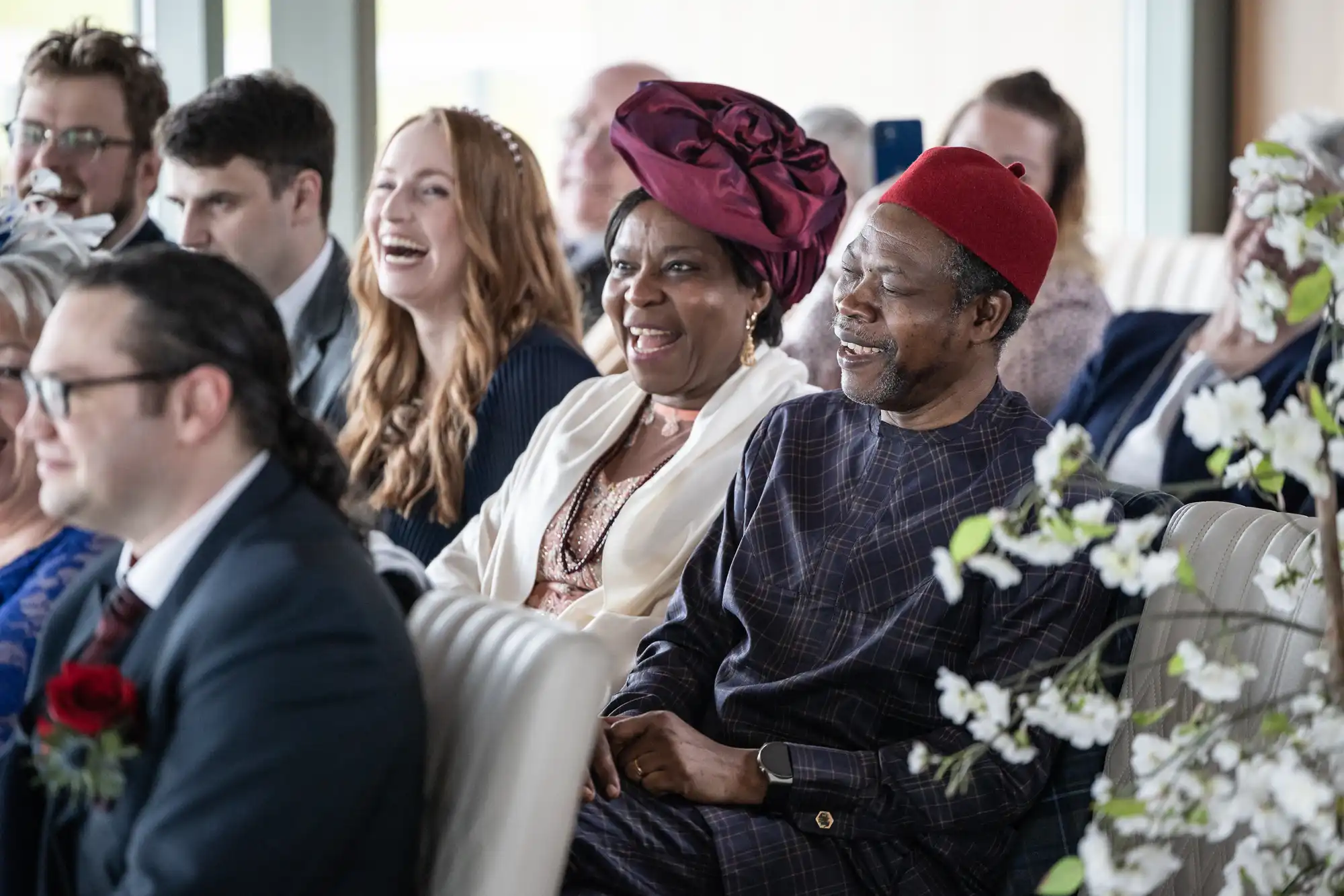 A diverse group of people sit in rows, smiling and laughing during an indoor event. In the foreground, a man and woman in colourful traditional African attire sit together, surrounded by others in formal clothing. White flowers decorate the right side of the image.