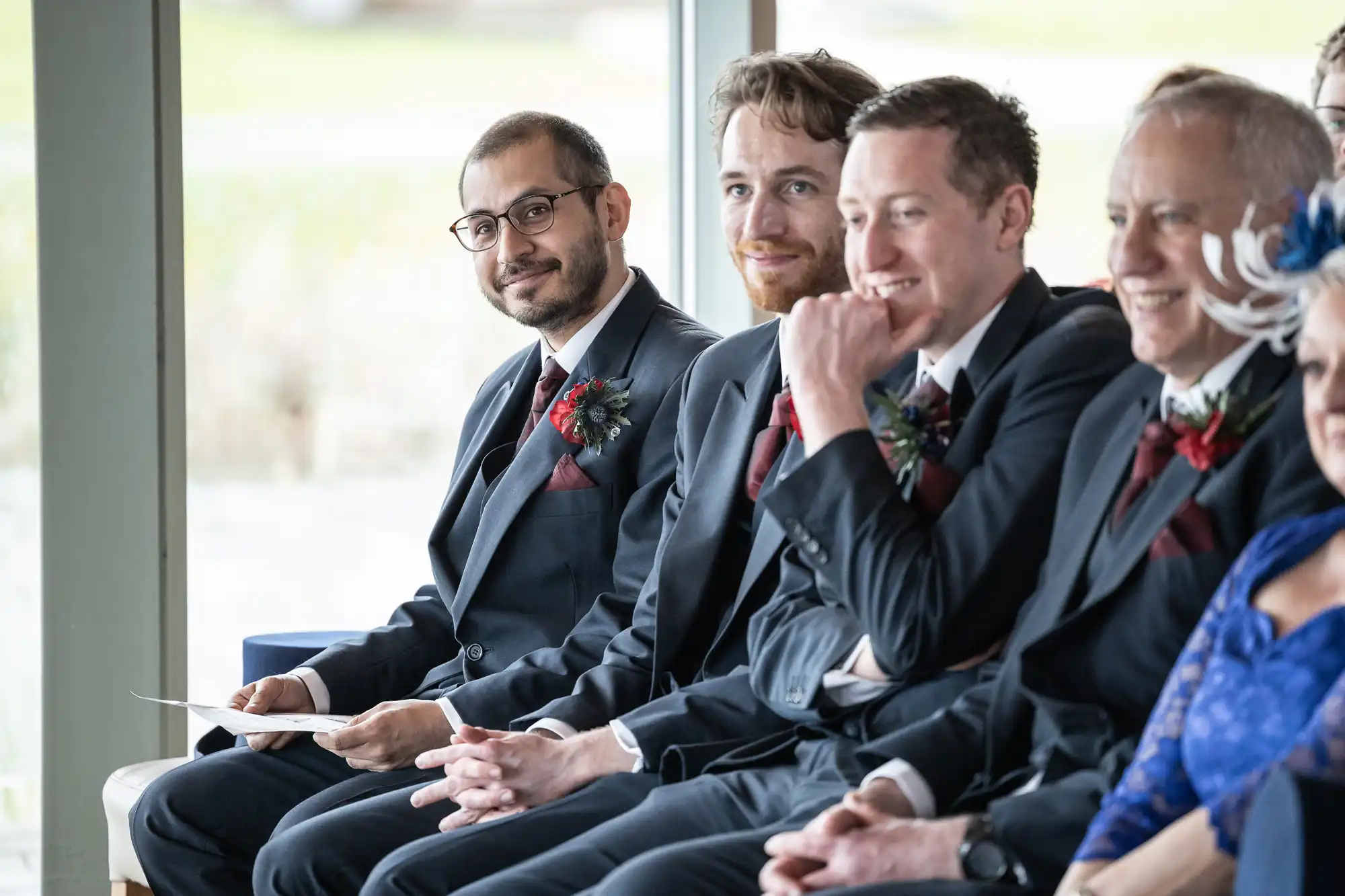 Four men in dark suits with buttonholes sit side by side at a wedding ceremony, smiling and looking forward. The man on the left holds paper, while the others chat and laugh. Light streams in from large windows behind them.