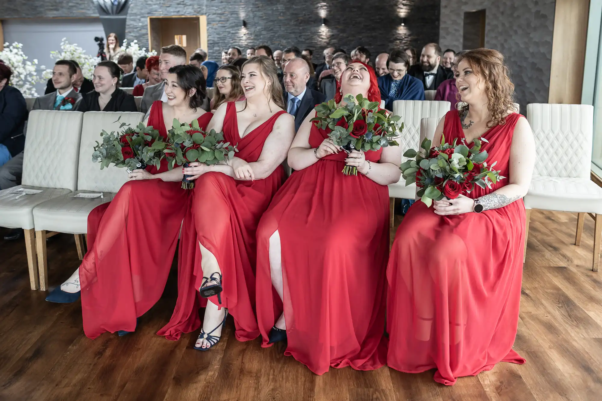 Four bridesmaids in matching red dresses sit on a white bench, laughing and holding bouquets of red and white flowers. Guests sit behind them in a modern, light-filled venue with wooden floors and stone walls. The atmosphere is joyful and candid.