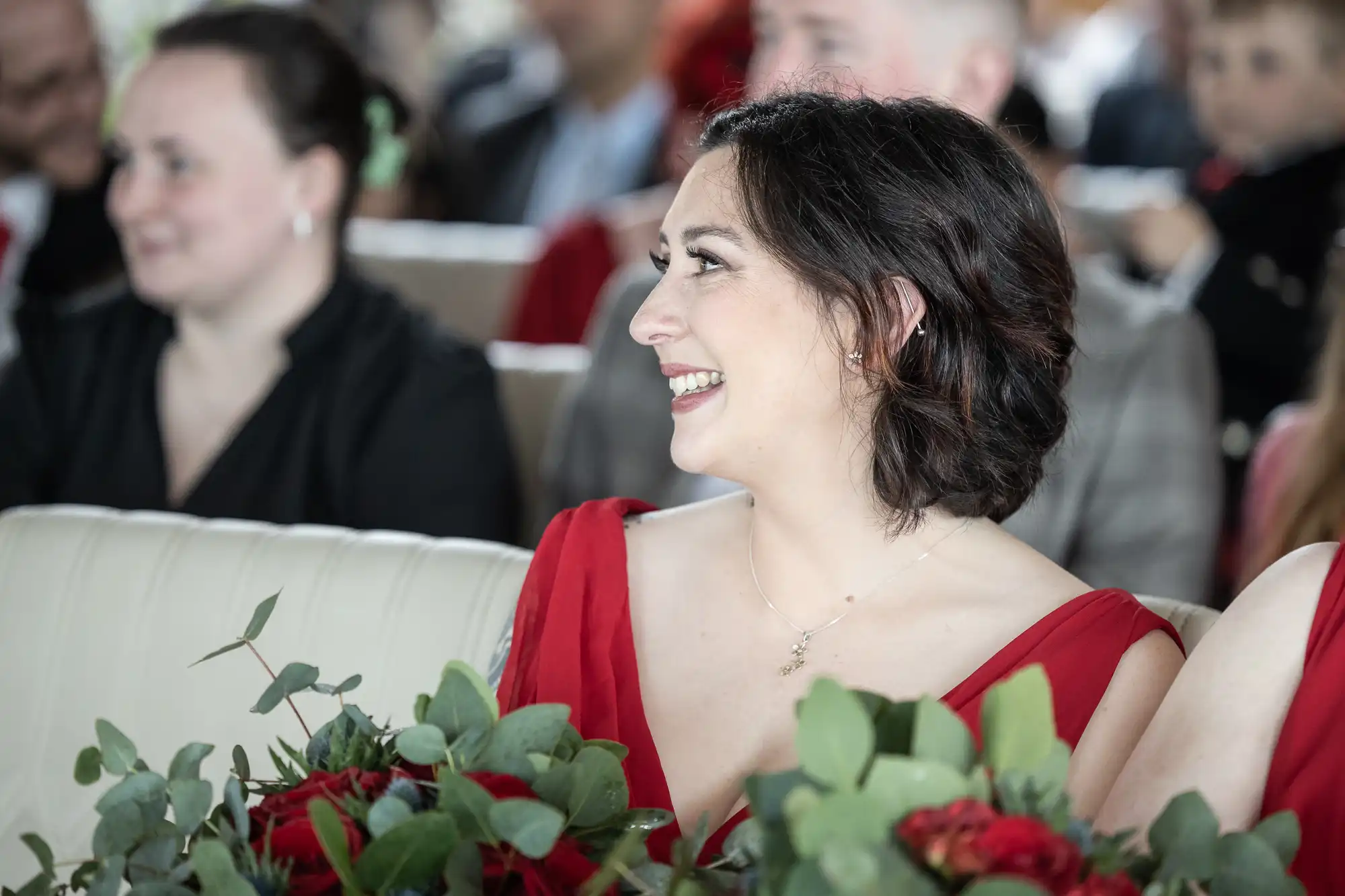 A woman with short dark hair, wearing a red dress and holding a bouquet of greenery and red flowers, smiles whilst seated indoors among a group of people. The background is softly blurred, highlighting her joyful expression.
