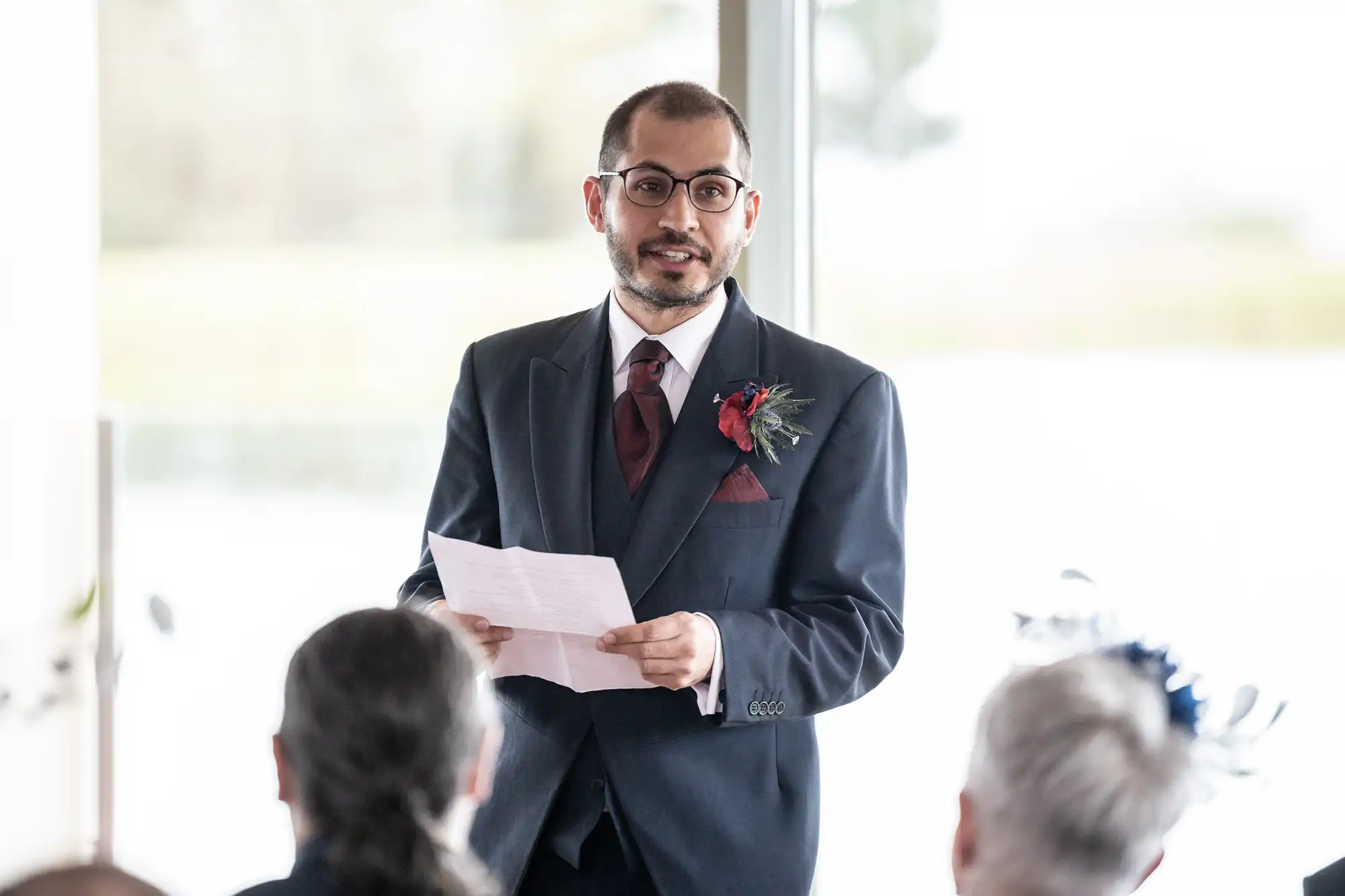 A man in a dark suit, maroon tie, and glasses stands indoors, holding a sheet of paper and speaking. He has a buttonhole on his lapel. Several people with grey or pinned-back hair sit facing him, their backs to the camera. Bright window light fills the background.