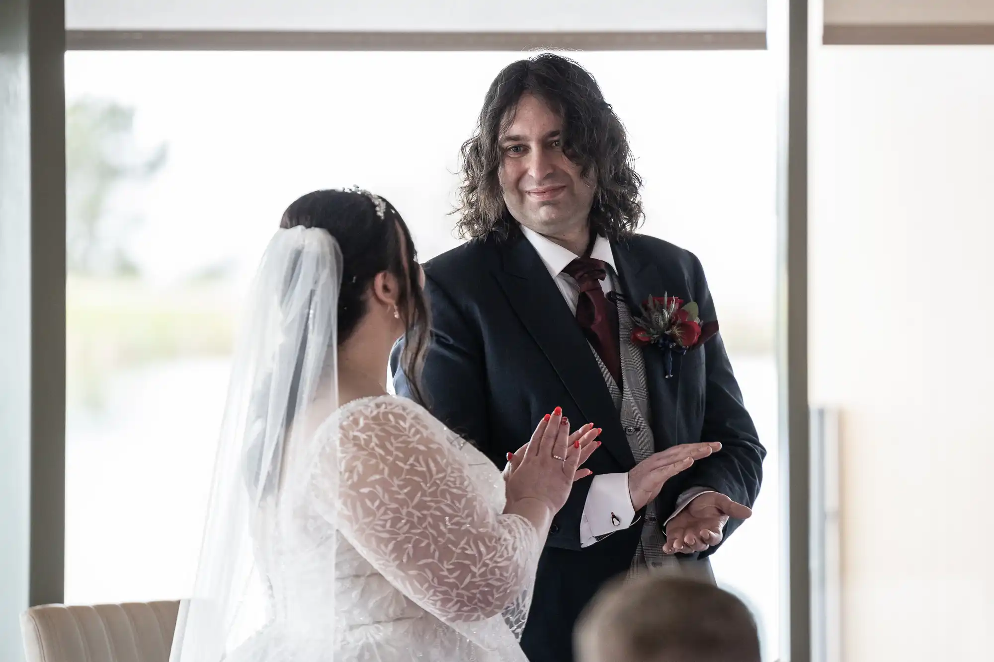 A bride in a white lace gown and veil faces a groom in a dark suit with a burgundy tie and buttonhole. The groom looks at her, smiling warmly, as she holds her hands together. They stand indoors near large windows on their wedding day.