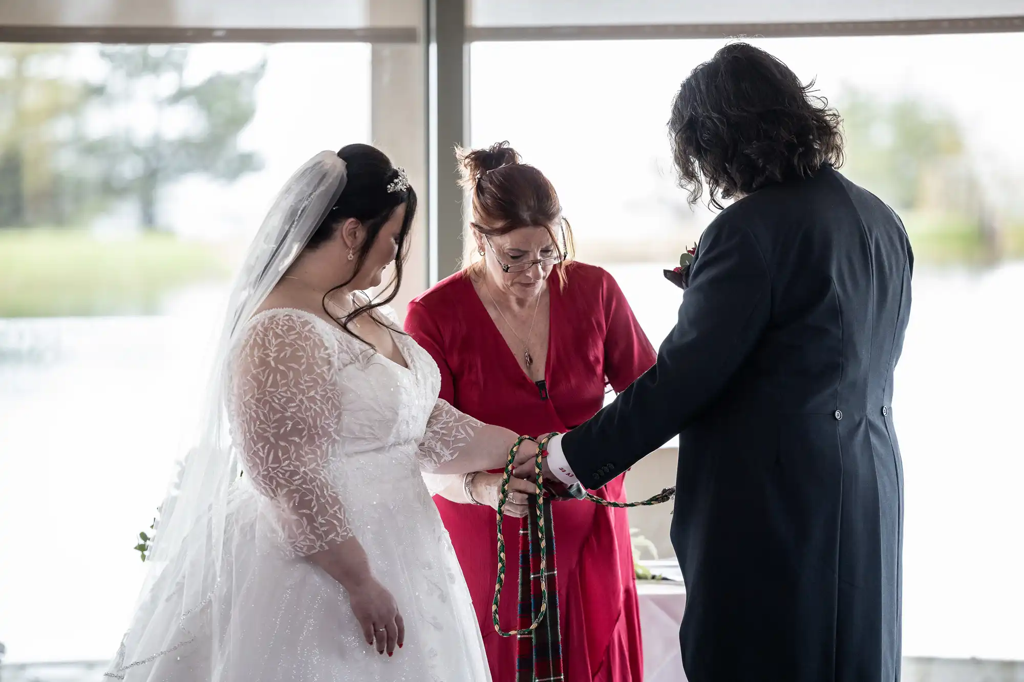 A bride in a white lace gown and veil and a groom in a dark suit stand holding hands, their hands wrapped in braided cords. A woman in a red dress officiates, looking down at their hands. Bright windows and a blurred outdoor scene are in the background.