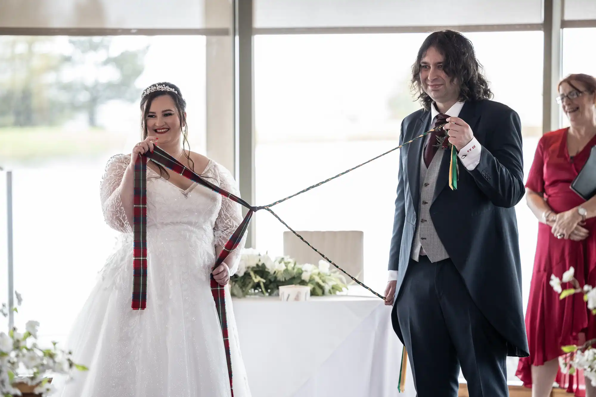 A bride in a white lace gown and a groom in a tuxedo smile as they hold tartan ribbons loosely tying their hands together during a handfasting ceremony. An officiant in a red dress stands nearby, and light streams in through large windows behind them.