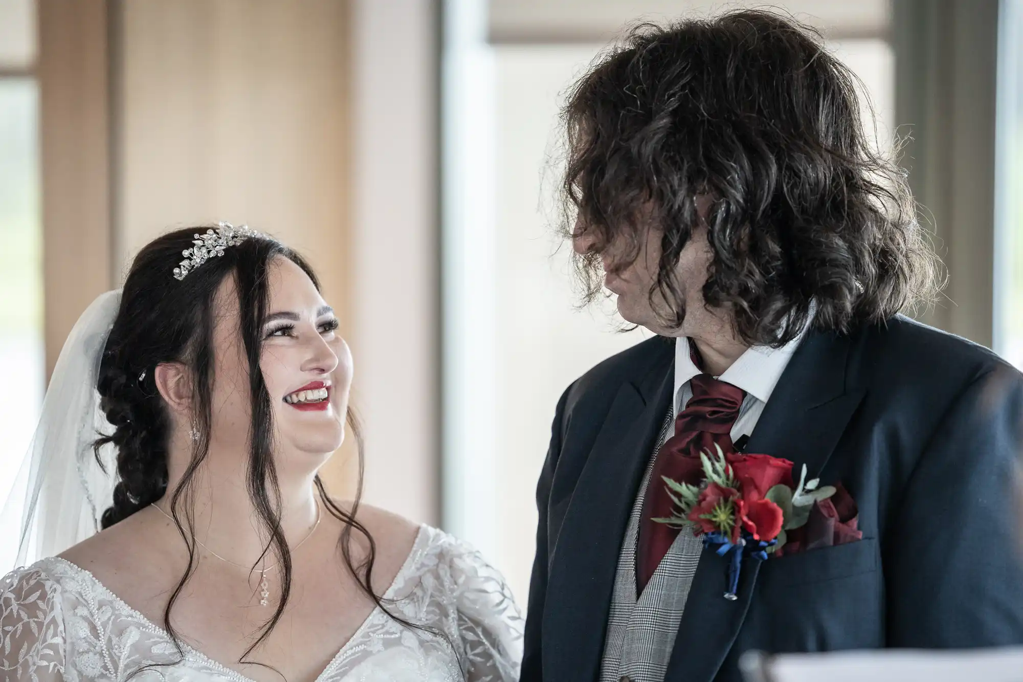 A bride in a white dress and veil smiles joyfully at the groom, who wears a black suit and maroon tie with a red buttonhole. They stand indoors, facing each other, sharing a happy moment on their wedding day.