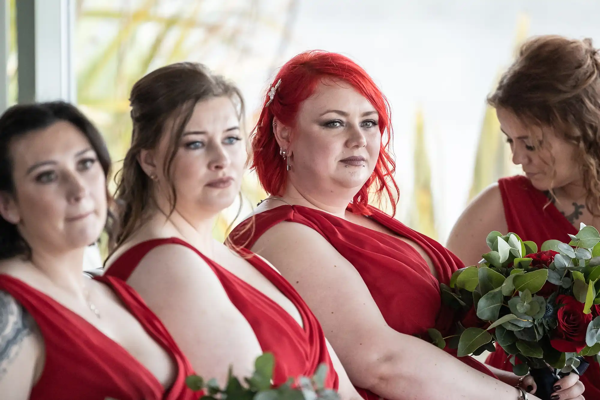 Four women stand together, wearing matching sleeveless red dresses and holding bouquets of green foliage and red flowers. Three women face forwards with serious expressions, whilst the fourth looks down at her bouquet. The background is softly blurred.