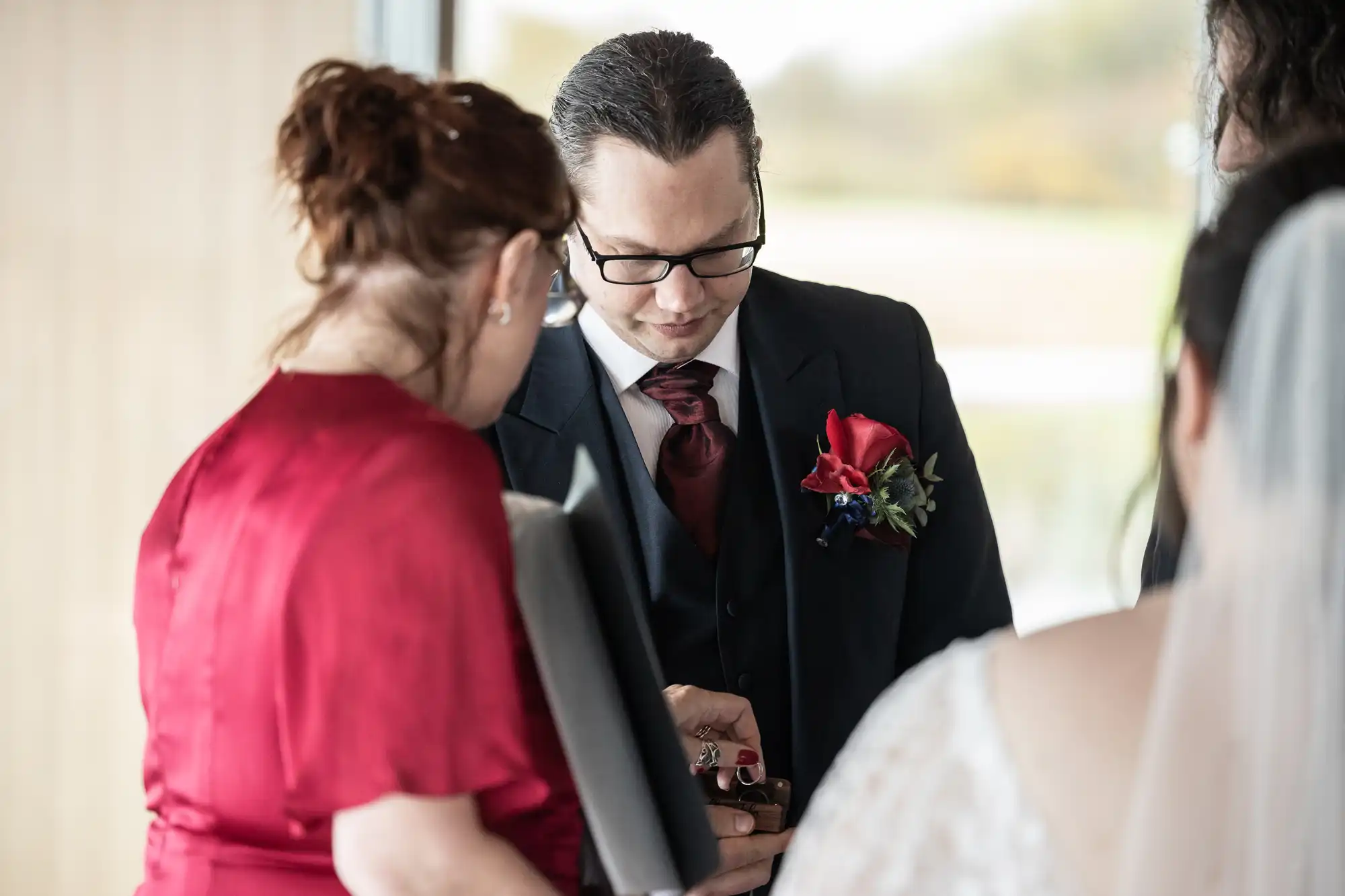 A man in a suit with a red tie and buttonhole looks down at an item held by a woman in a red dress. Another person in a white lace dress, likely a bride, stands nearby. The indoor setting is softly lit with a blurred window view behind them.