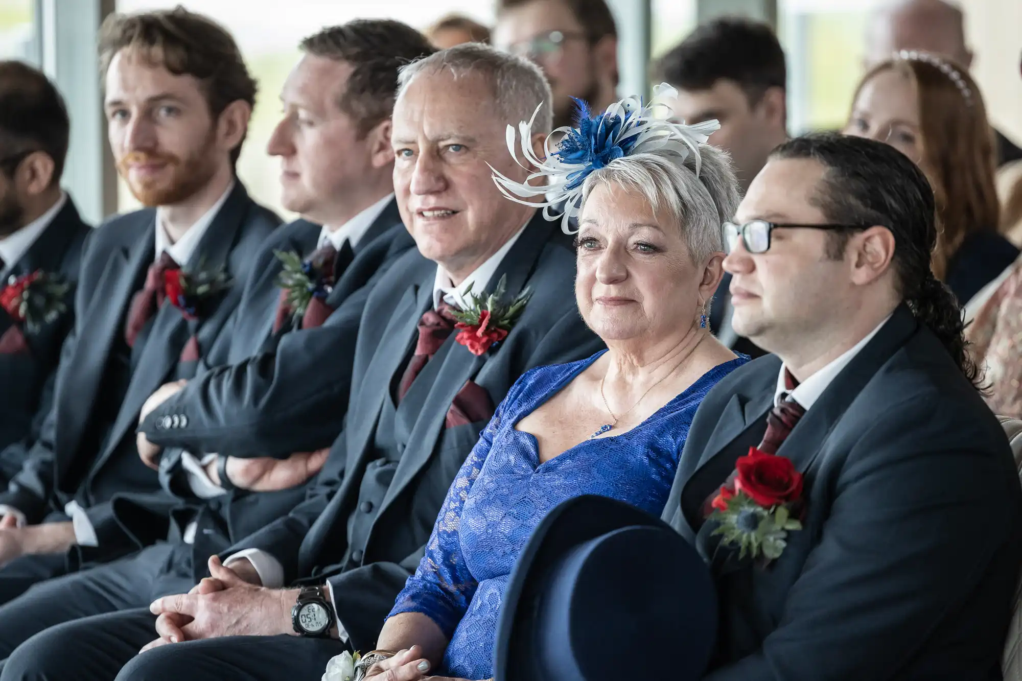 A row of elegantly dressed wedding guests sit indoors, mostly men in dark suits with burgundy ties and buttonholes. A smiling woman in a blue lace dress and feathered fascinator sits among them, holding a navy hat. The group looks forward, attentive and happy.