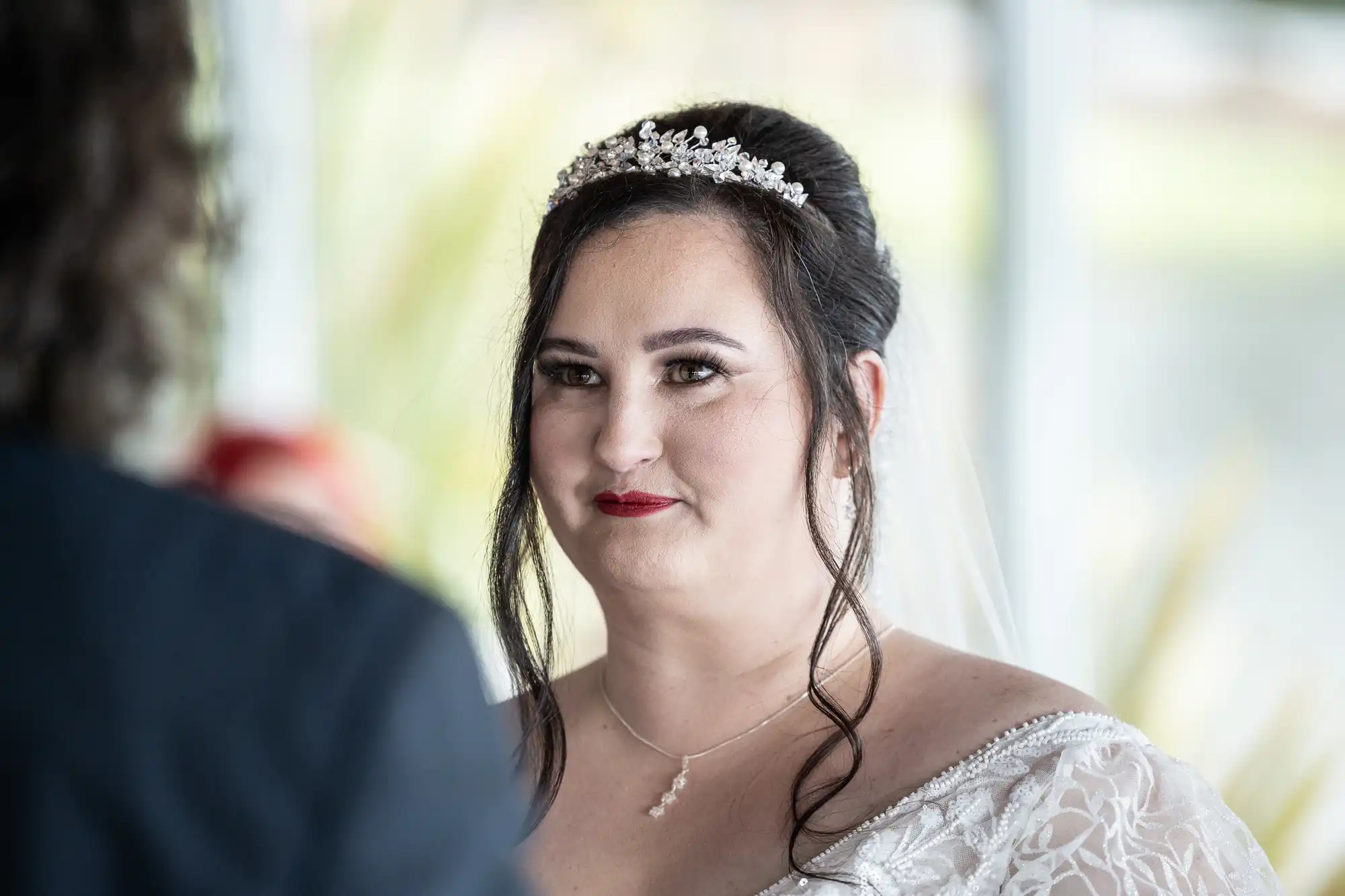 A bride with dark hair in an updo, wearing a jewelled tiara, white lace wedding dress, and veil, looks forward with a gentle smile. She has natural make-up and a pendant necklace. The background is softly blurred with green and light tones.