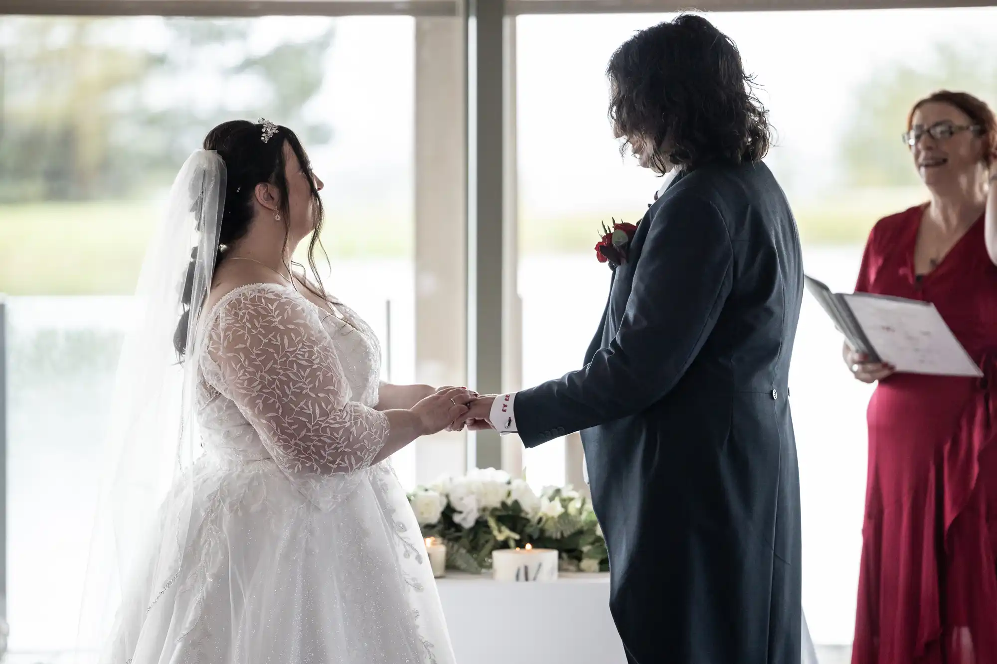 A bride in a white dress and veil holds hands with a groom in a dark suit during a wedding ceremony. An officiant in a red dress stands nearby, smiling and holding papers. Soft natural light streams in through large windows behind them.