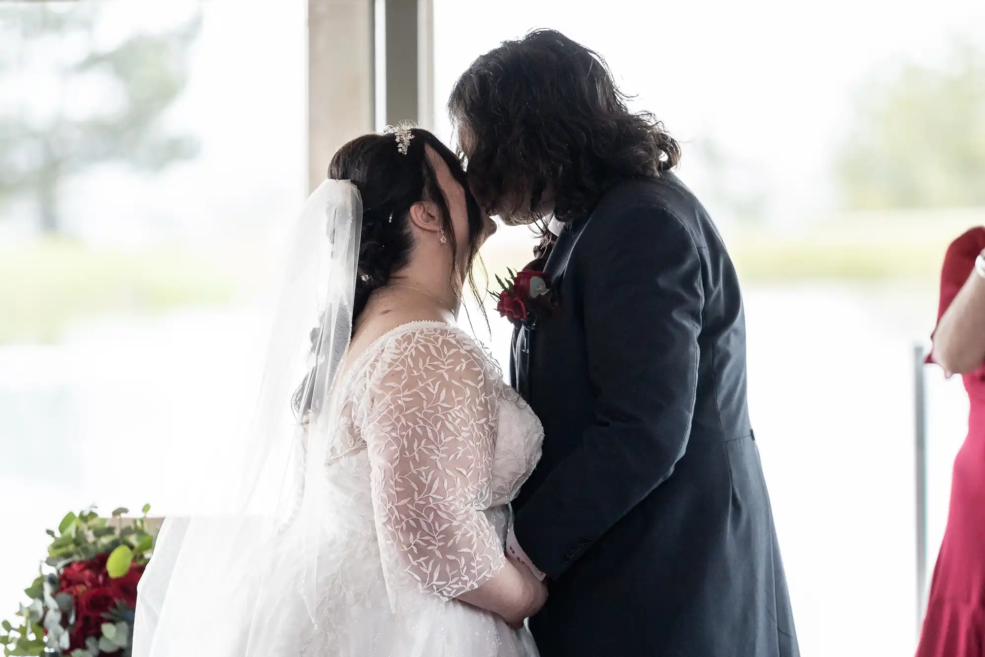 A bride in a white lace gown and veil kisses a groom in a navy suit with a red buttonhole. They stand indoors near a window, with natural light illuminating them. A bouquet with red and green flowers is visible on the left.
