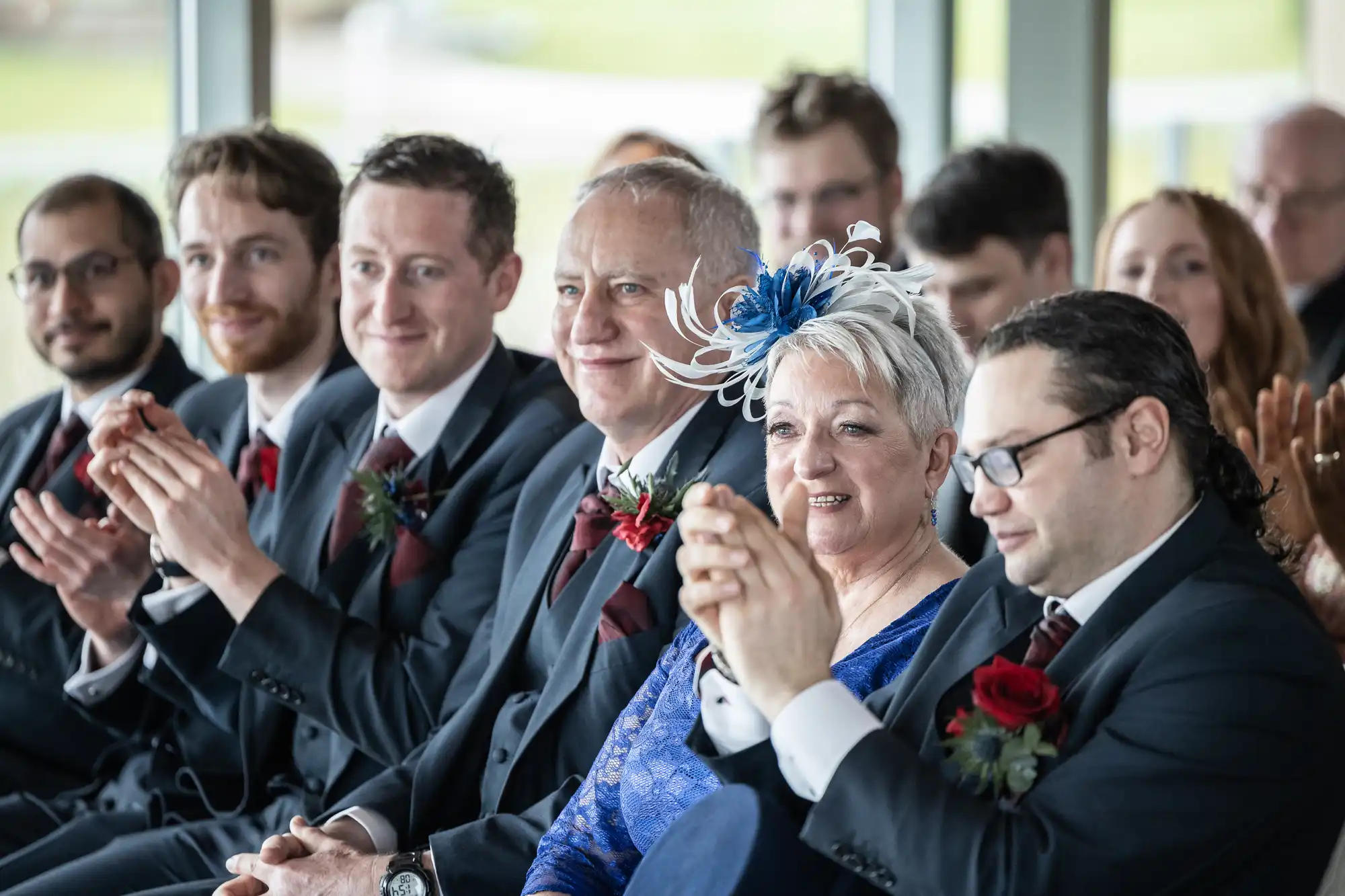 A group of people in formal attire sit in rows, smiling and clapping. The woman in front wears a blue lace dress and a decorative fascinator. Men beside her wear dark suits with red ties and buttonholes, suggesting they are attending a wedding or formal event.