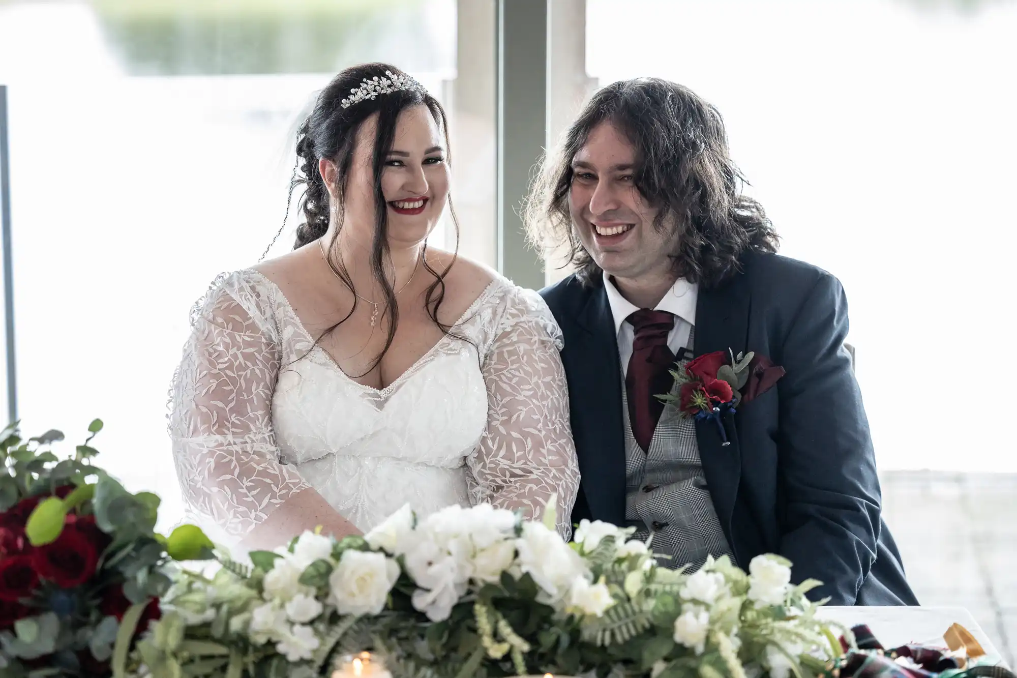 A bride in a lace-sleeved white dress and a groom in a dark suit with a maroon tie sit smiling together at a table decorated with white and red flowers and greenery, with a bright windowed background behind them.