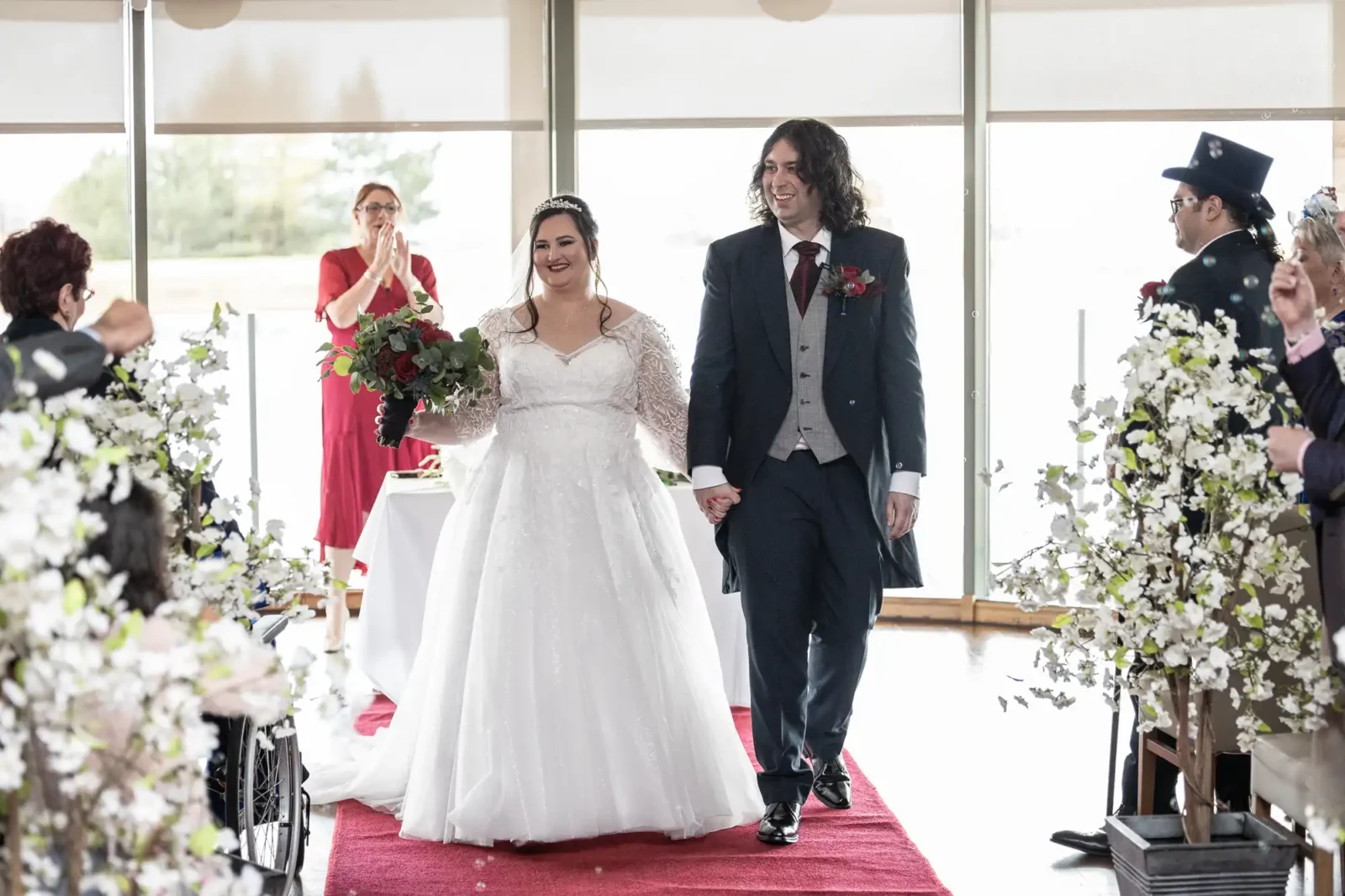 A smiling bride in a white lace dress and a groom in a black suit walk hand in hand down a red aisle, surrounded by white floral arrangements and applauding guests at an indoor wedding ceremony.