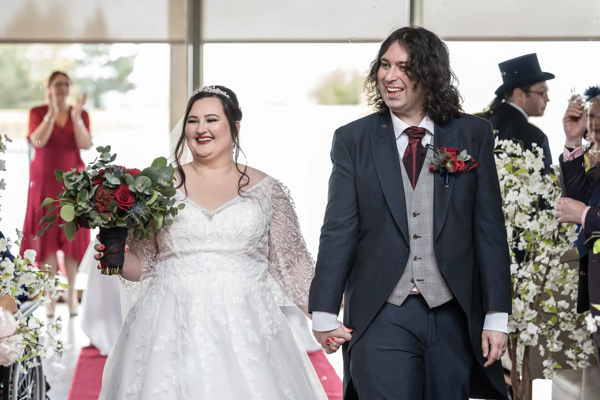 A smiling bride in a white gown holds a bouquet of red and white flowers, walking hand-in-hand with a groom in a dark suit and red buttonhole. They are indoors, surrounded by cheering guests and floral decorations, celebrating their wedding.