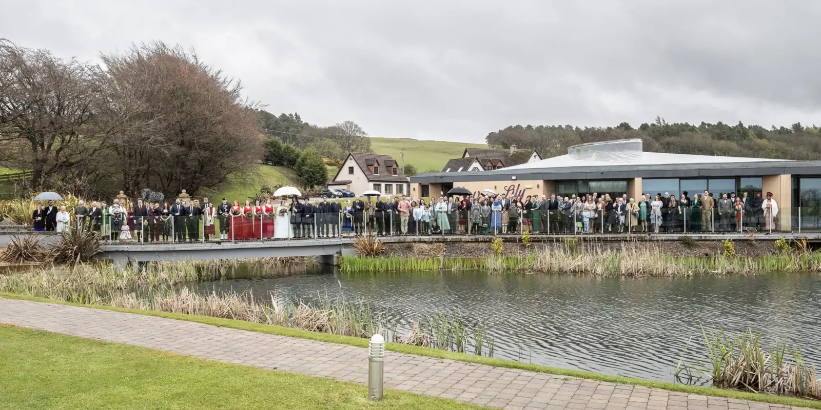 A large group of people dressed formally stand on a long bridge over a pond, posing for a photo. The background has a modern building, houses, grassy hills, and cloudy sky. Trees and reeds border the water, with a paved path in the foreground.