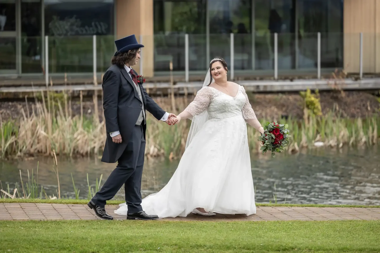 A bride in a white dress holds a bouquet of red flowers and smiles whilst walking hand-in-hand with a groom in a dark suit and top hat along a path by a pond, with reeds and a modern building in the background.