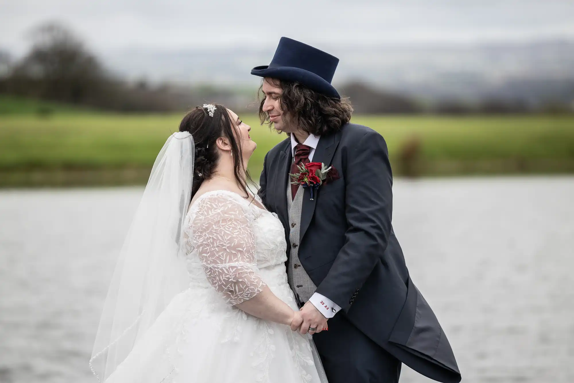 A bride in a white lace wedding dress and veil holds hands and smiles at a groom in a dark suit with a top hat. They stand outdoors by a lake, with green grass and trees in the blurred background.