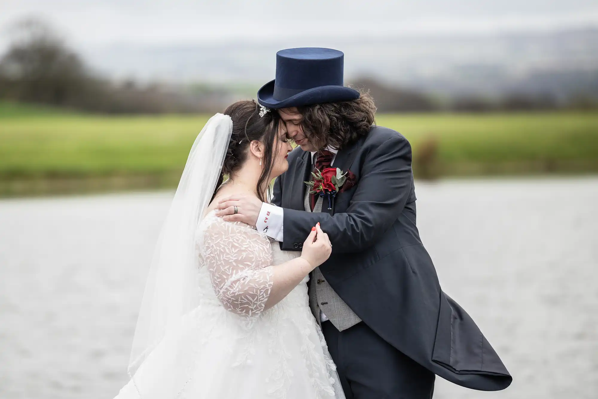 A bride in a white, lace-sleeved wedding dress and veil embraces a groom in a dark suit, grey waistcoat, and blue top hat. They smile and press their foreheads together by a lake, with green fields and cloudy sky in the background.