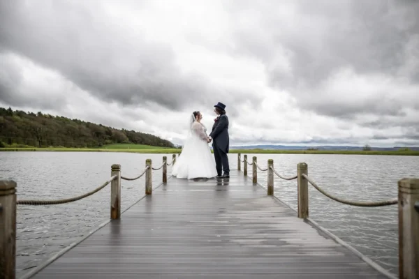 A bride and groom stand facing each other on a wooden jetty over a lake, holding hands. The bride wears a white gown and veil; the groom wears a dark suit and top hat. The sky is cloudy, and green hills are visible in the background.