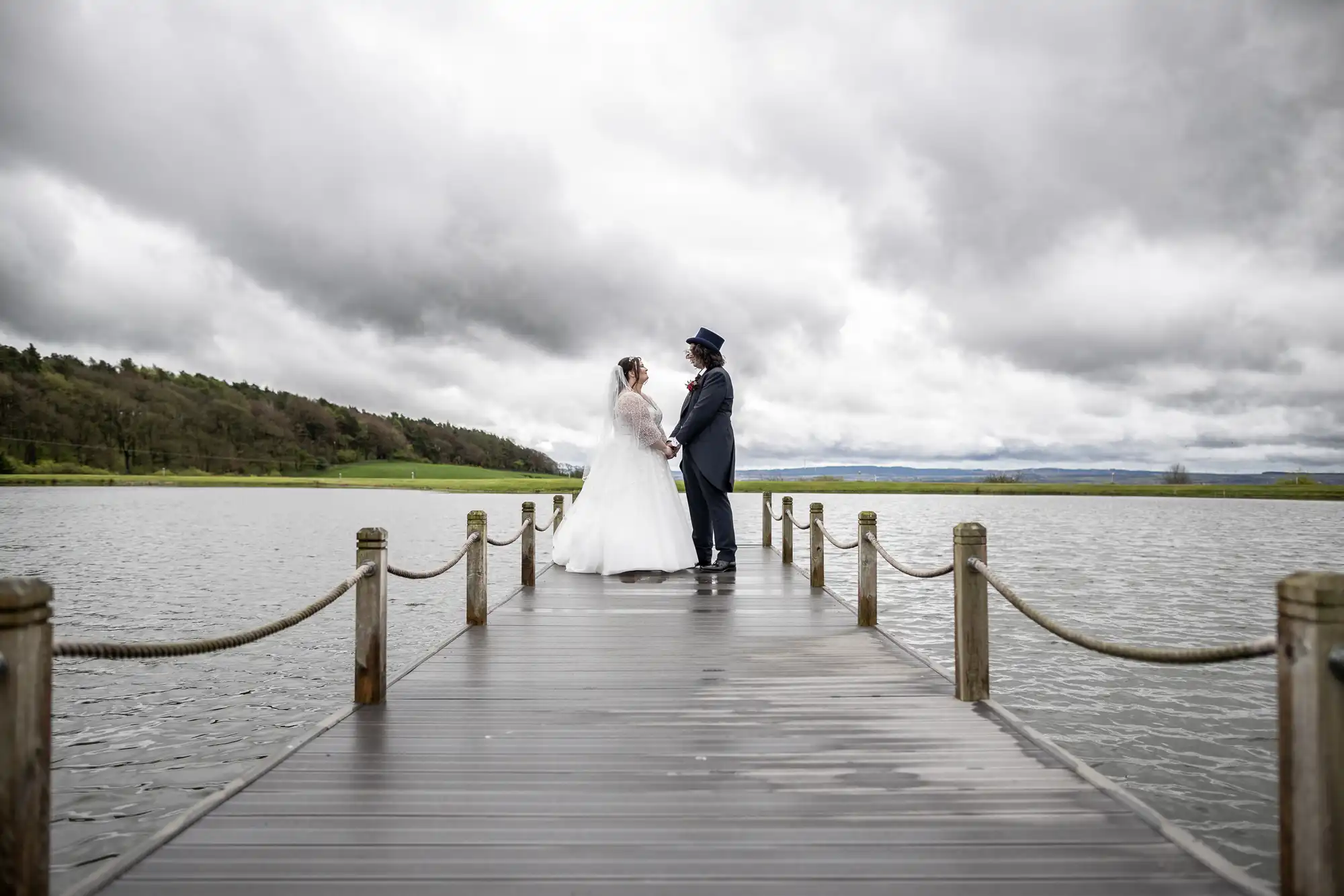 A bride and groom stand facing each other on a wooden jetty over a lake, holding hands. The bride wears a white gown and veil; the groom wears a dark suit and top hat. The sky is cloudy, and green hills are visible in the background.