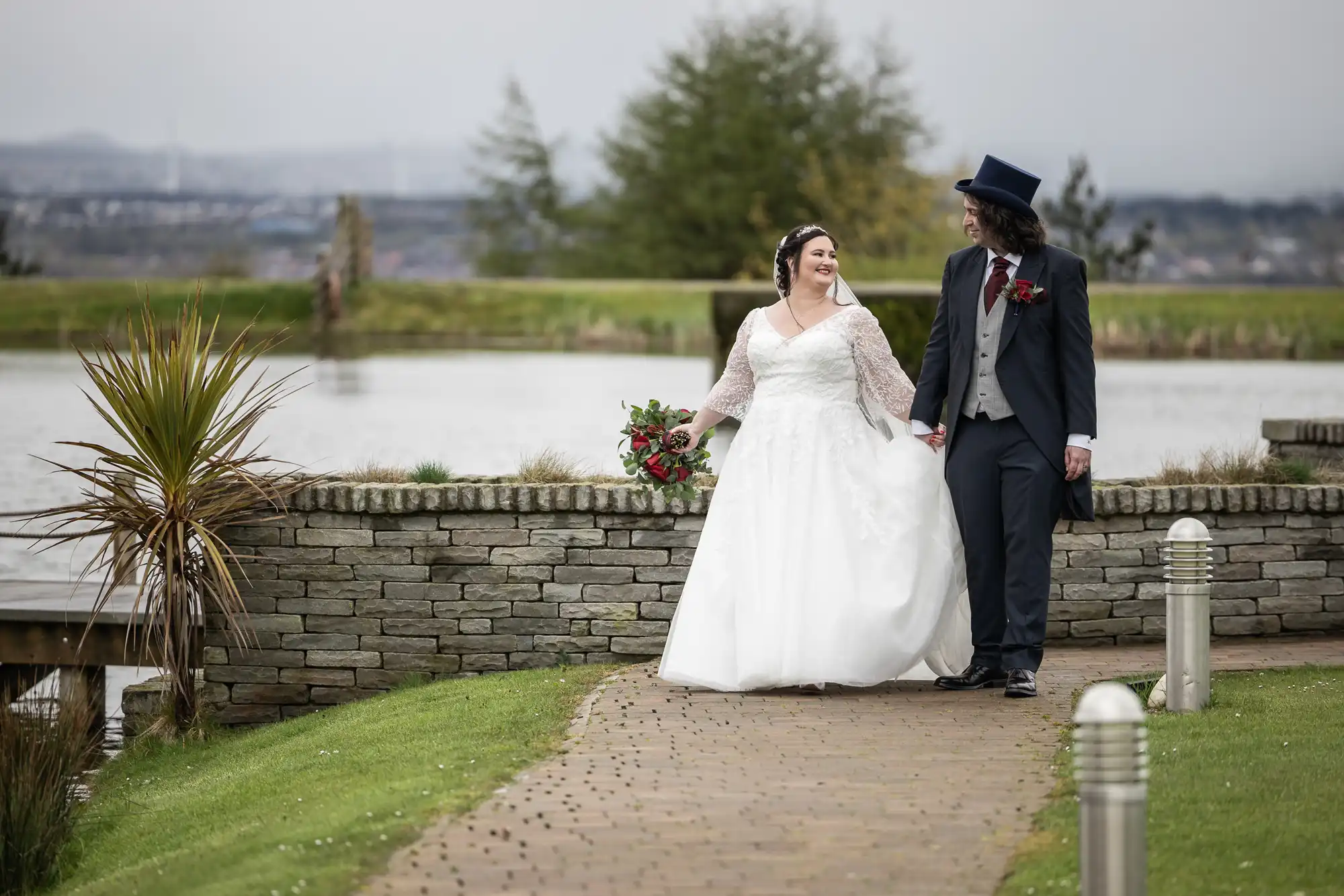 A bride in a white dress and a groom in a dark suit with a top hat walk hand in hand on a path by a lake, both smiling. The bride holds a red bouquet. Green grass, a stone wall, and trees surround them; water and hills are in the background.