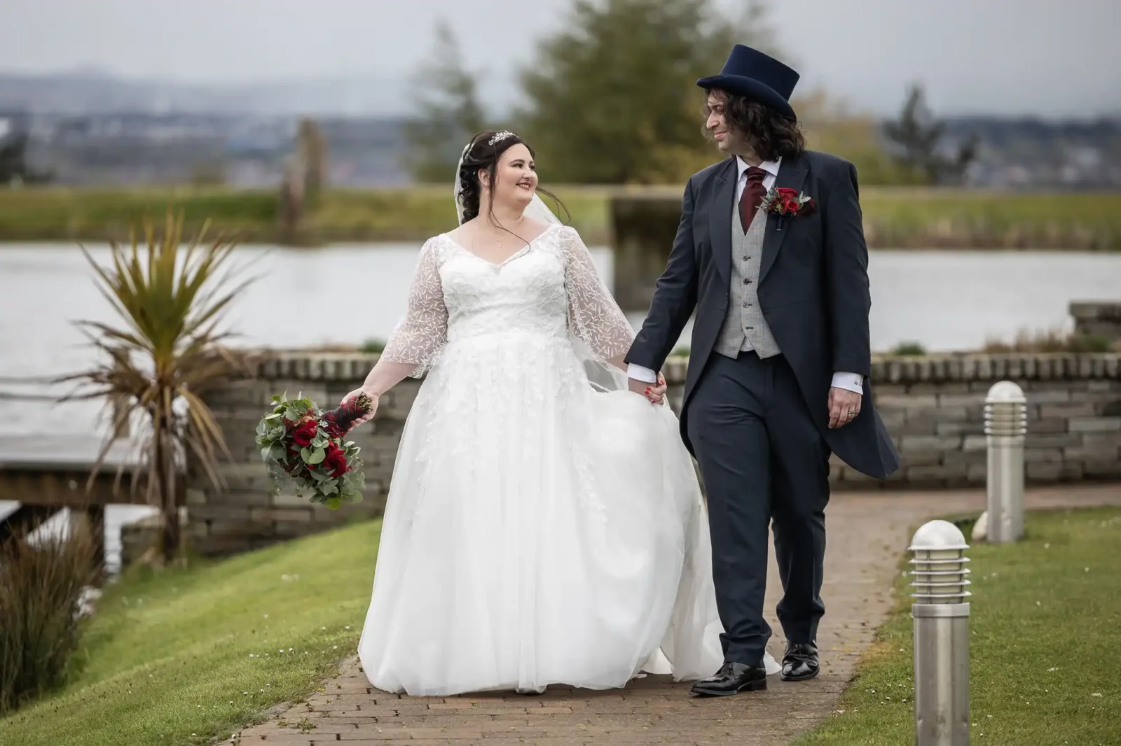 The Vu wedding photos image of a bride in a white dress and veil holds a bouquet of red flowers and walks hand in hand with a groom in a dark suit, top hat, and red buttonhole on a garden path beside a lake, both smiling at each other.