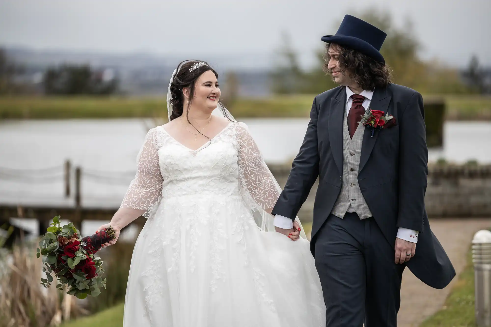 A bride in a white lace wedding dress holds a red bouquet and smiles at a groom in a navy suit, grey waistcoat, red tie, and top hat. They walk hand-in-hand outdoors near water, with blurred trees and fencing in the background.