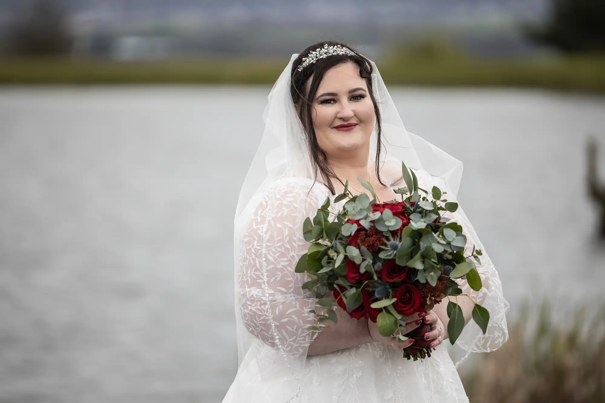 A bride in a white lace dress and veil stands by a lake, smiling and holding a bouquet of red roses and foliage. She wears a tiara and dark lipstick. The background is out of focus, with water and greenery visible.
