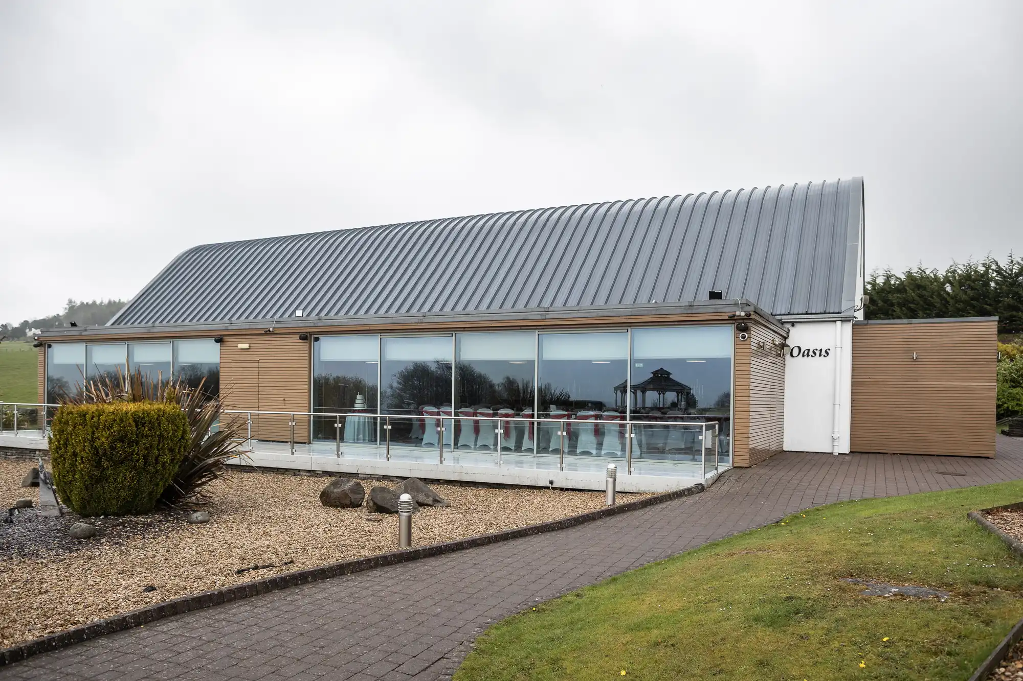 A modern, single-storey building with a sloped grey metal roof and large glass windows labelled "Oasis." The building is surrounded by landscaped gravel, shrubs, and a paved walkway, with grassy fields and trees in the background under a cloudy sky.