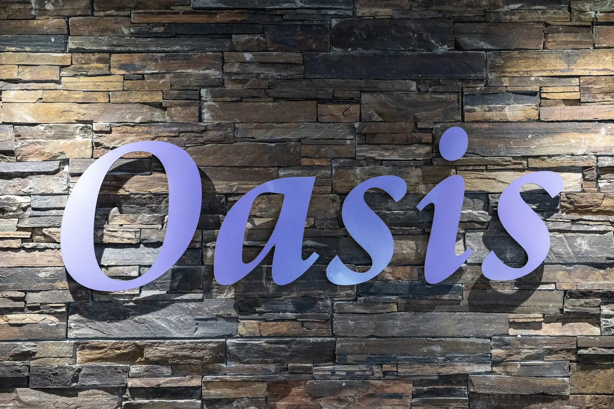 Large, purple letters spell "Oasis" on a wall made of horizontal, multicoloured stone bricks. The lighting creates soft shadows behind the text, highlighting the textured stone surface in shades of brown, grey, and tan.