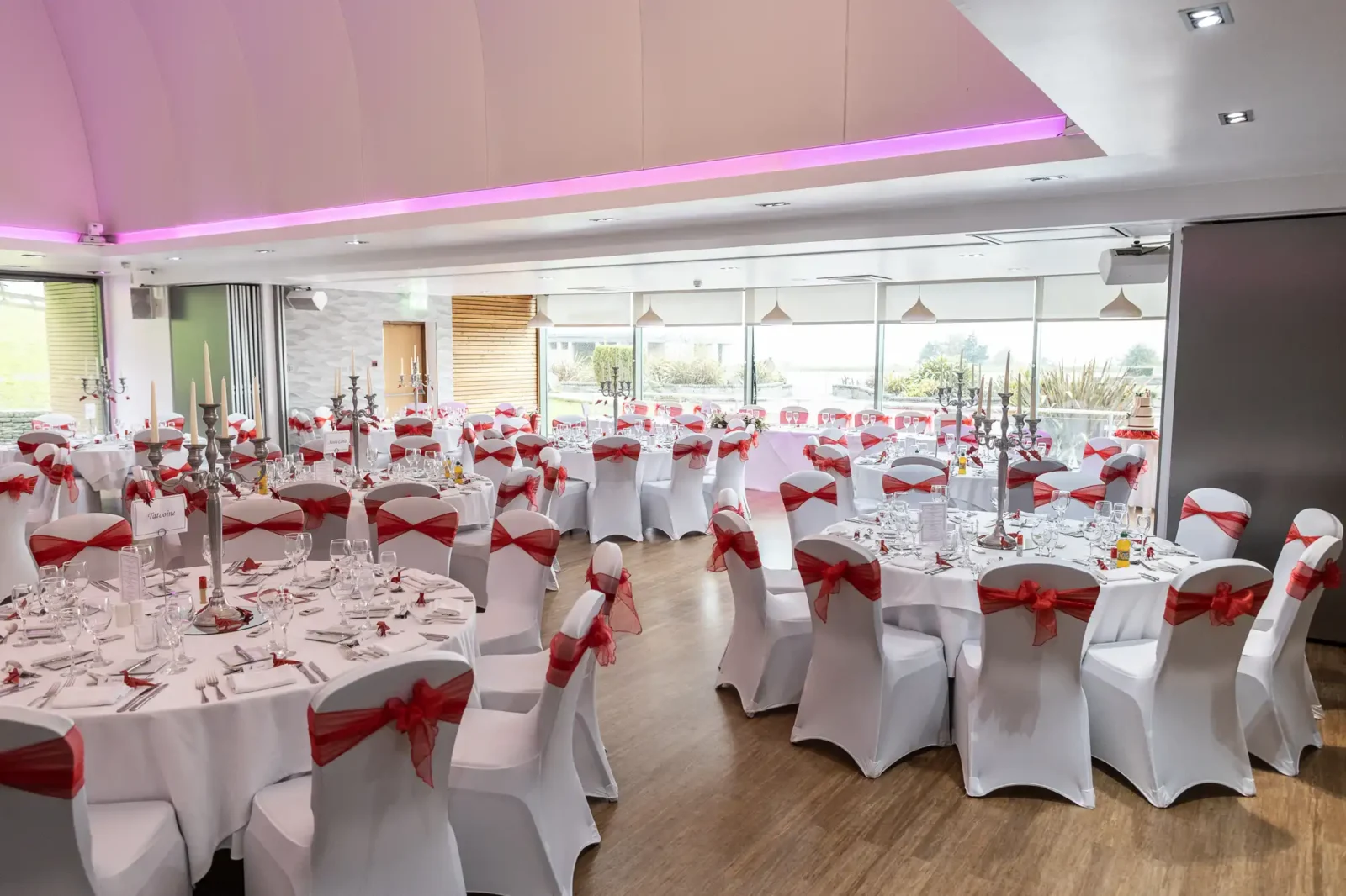 A bright banqueting hall with large windows and round tables set for an event. Each table is covered with a white cloth and set with glassware and cutlery. Chairs have white covers and red sashes tied in bows. Pink lighting accents the ceiling.