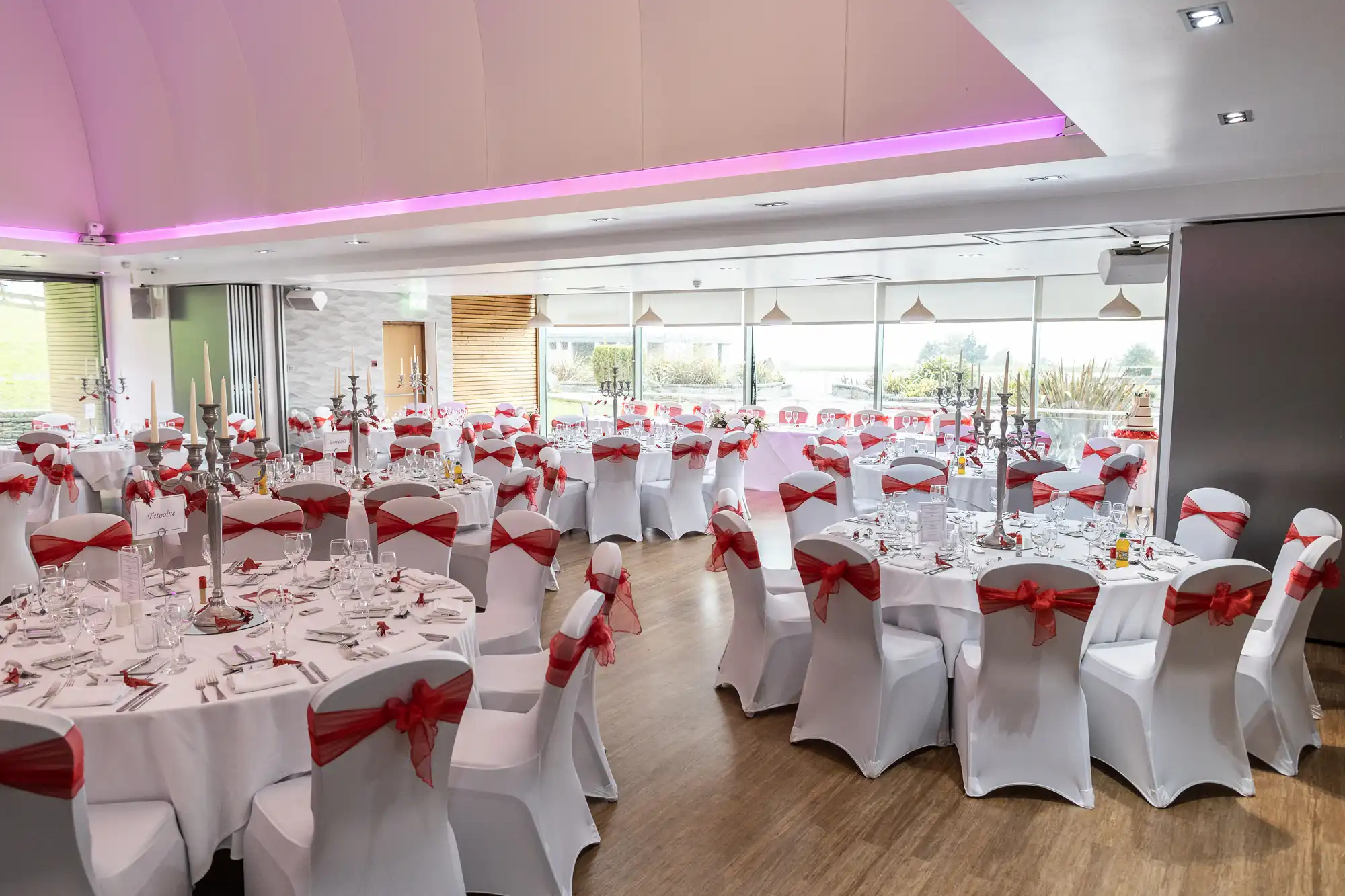 A bright banqueting hall with large windows and round tables set for an event. Each table is covered with a white cloth and set with glassware and cutlery. Chairs have white covers and red sashes tied in bows. Pink lighting accents the ceiling.