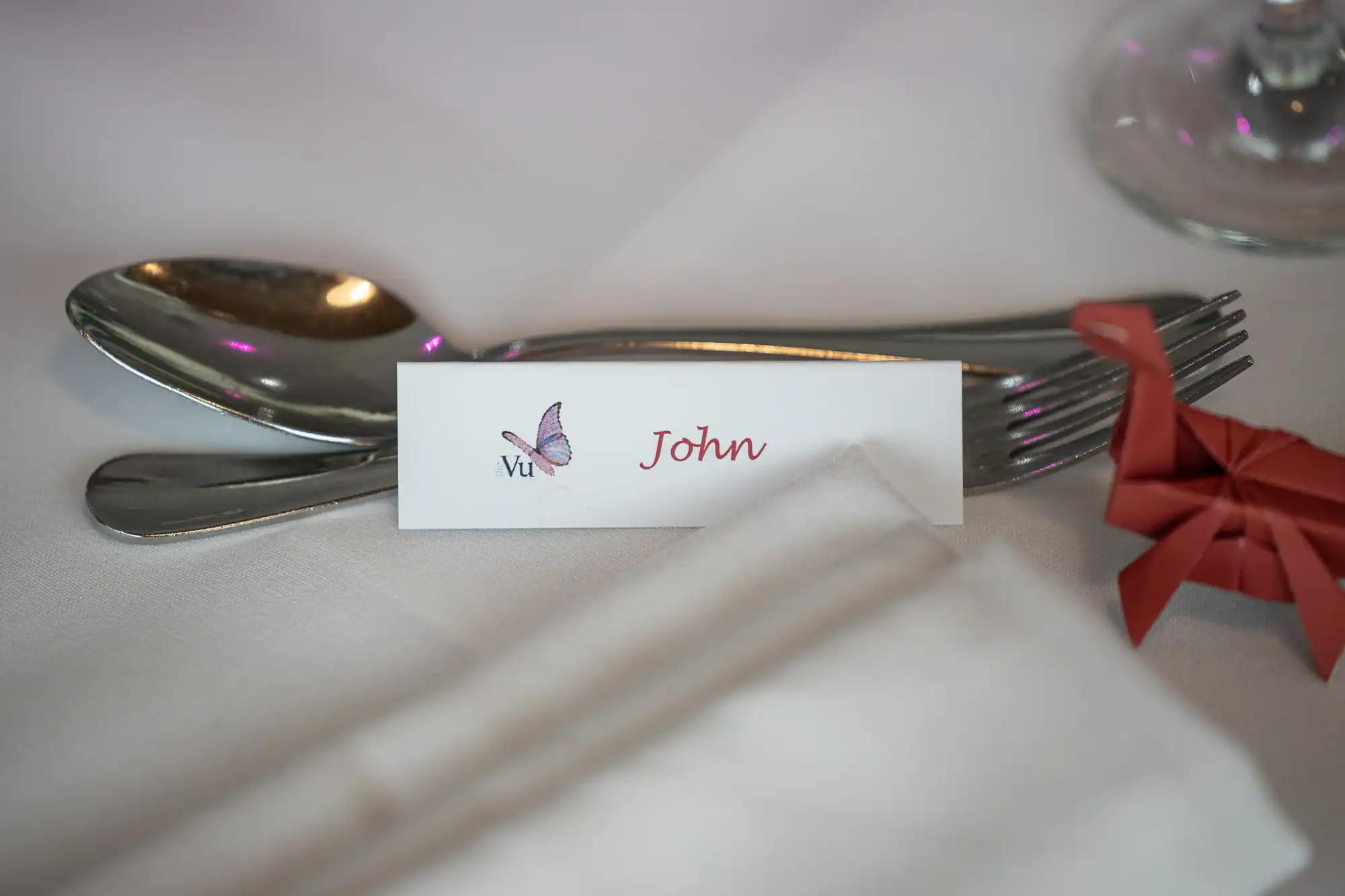 A place setting with a spoon and fork on a white tablecloth. In front is a name card reading "Vu John" with a purple butterfly illustration. An orange origami decoration is to the right, and a white serviette is in the foreground.