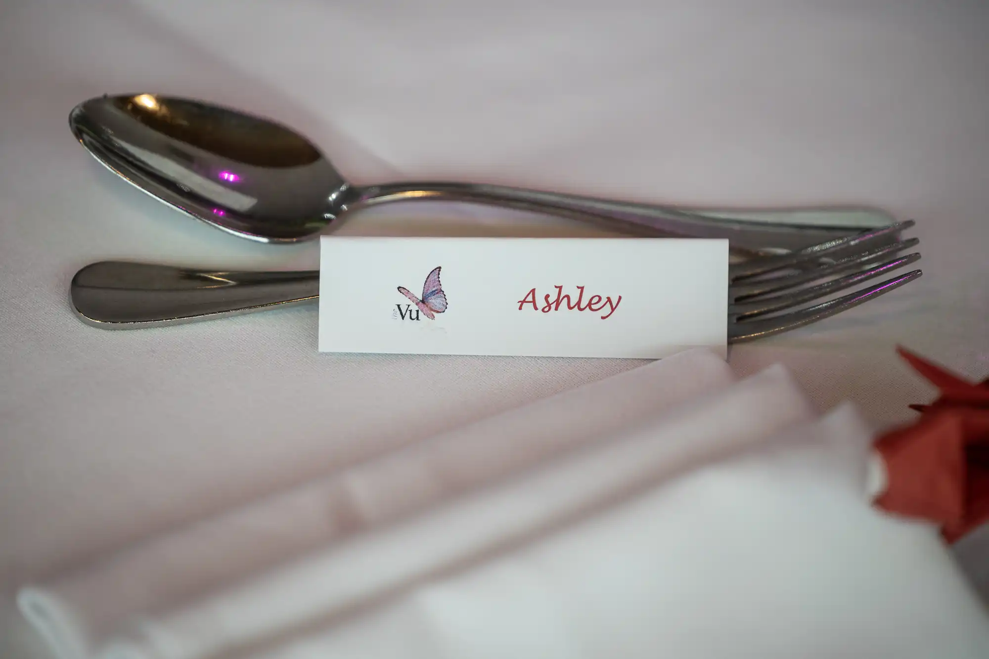 A close-up of a table setting with a folded white name card reading "Ashley" in red script, next to a purple butterfly illustration and “Vu.” Behind the card are a spoon and fork placed on a white tablecloth, with a white serviette in the foreground.