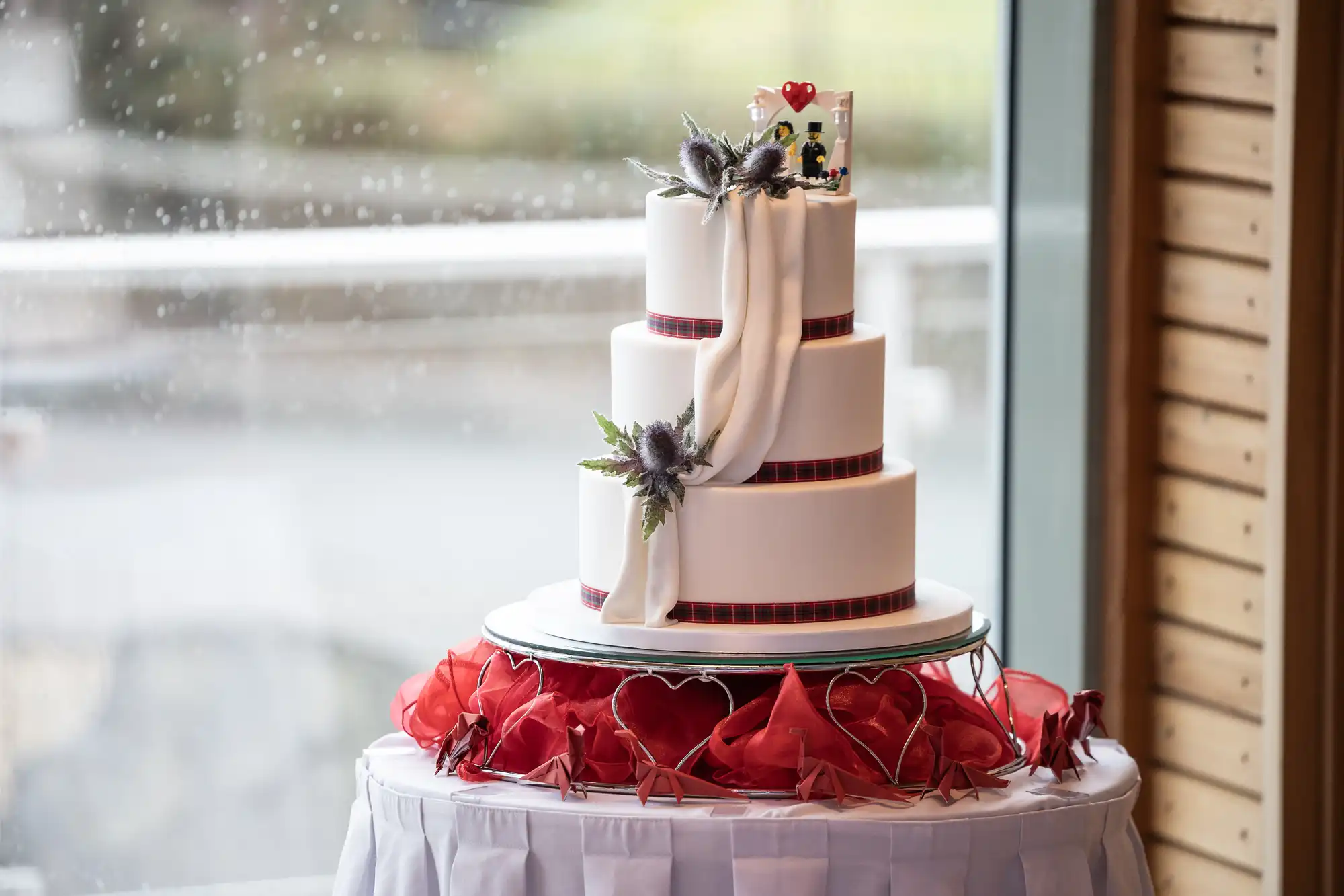 A three-tier white wedding cake with burgundy ribbon borders, decorated with a white drape, thistle flowers, and a small bride and groom topper. The cake sits on a round table adorned with red fabric and heart shapes, next to a window on a rainy day.