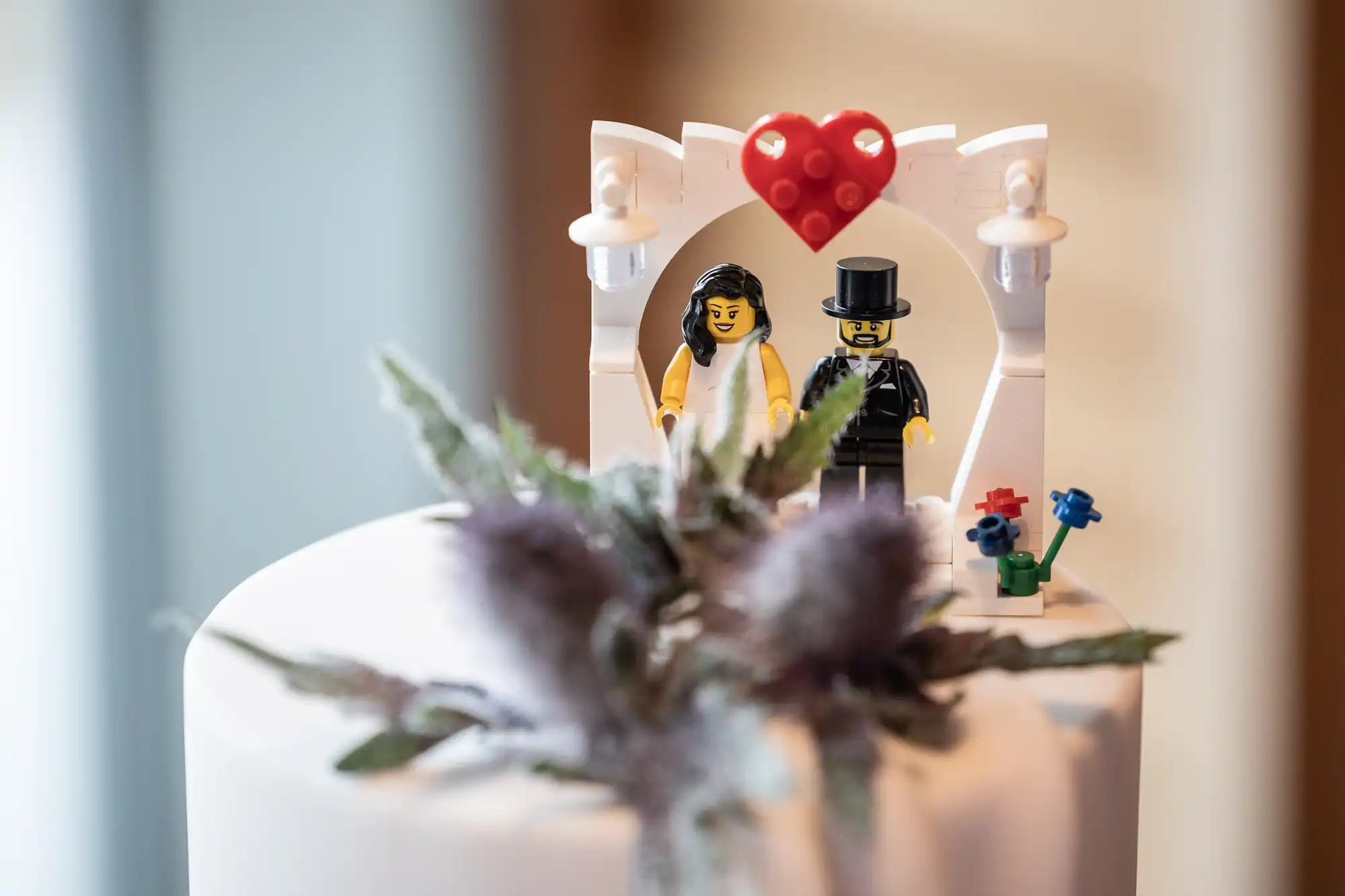 A close-up of a wedding cake topper made from LEGO figures of a bride in white and a groom in a black suit and top hat, standing under a white arch with a red heart. Blurred flowers decorate the cake in the foreground.