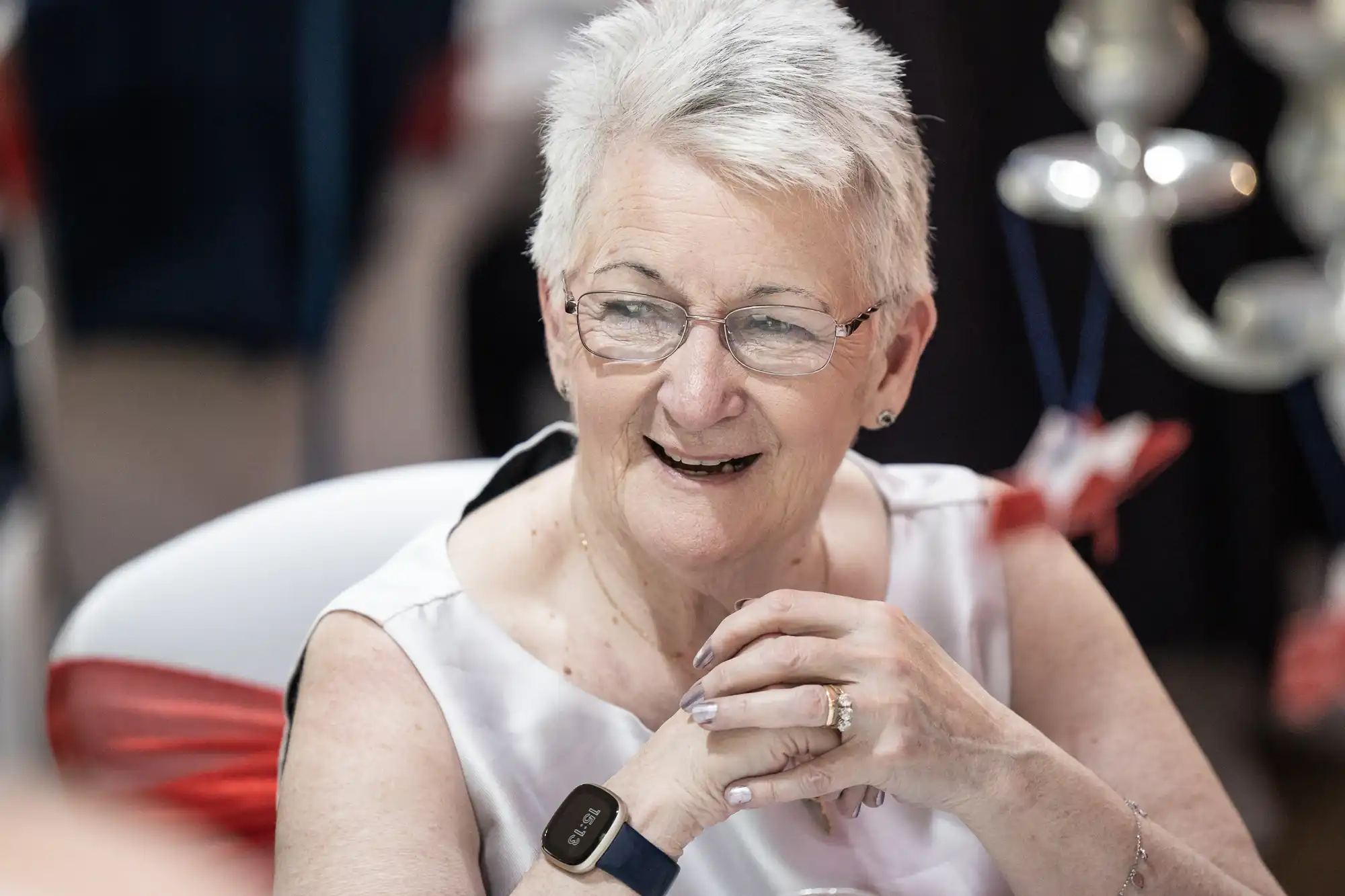 An older woman with short white hair and glasses smiles while seated at a table. She wears a sleeveless white top and a black smartwatch, with her hands clasped. The background is softly blurred with hints of red decorations.